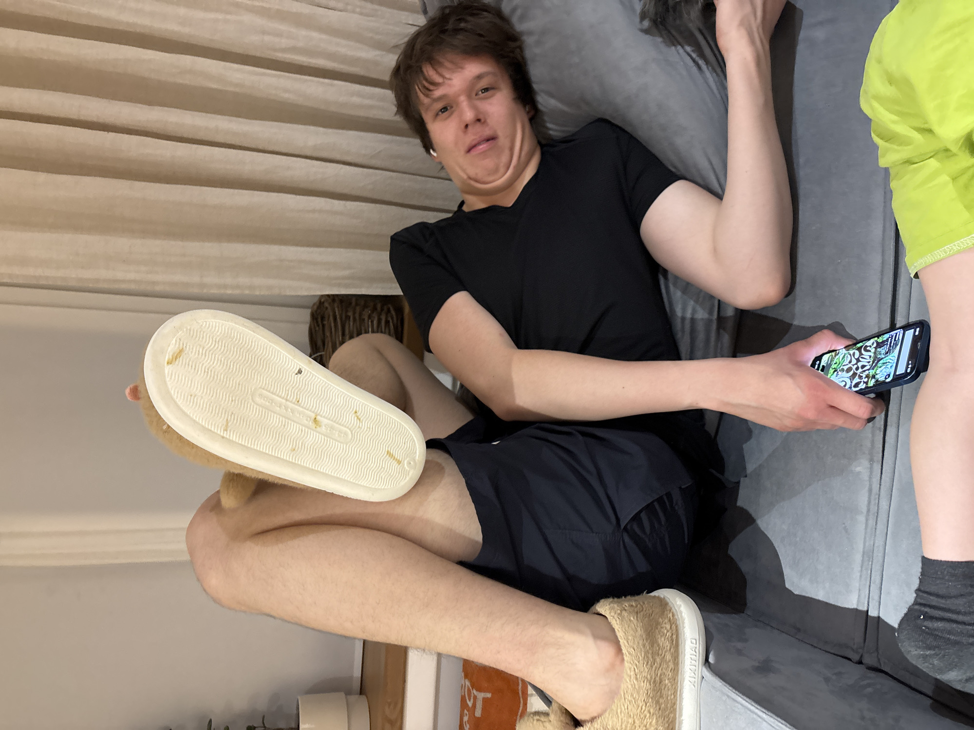The image depicts an indoor scene featuring two individuals in a residential setting. The specific location within Chaikovsky, Russia, is not identifiable from the visual evidence.
A male individual, appearing to be a young adult, is positioned supine on a grey upholstered surface, likely a couch or bed. He wears a black short-sleeved t-shirt and white wireless earbuds in both ears. His head is turned towards the viewer, and his facial muscles are contracted, creating a visible double chin.
Portions of a second individual are visible in the foreground. This individual's left leg, clad in a black ribbed sock and neon green shorts, is in the lower left. An elevated foot, wearing a light brown furry slipper with a white sole, is prominently displayed in the upper left-center. The slipper's side bears the inscription "XIMILIAD." The second individual's hand, partially visible in the lower right, holds a dark-colored smartphone, displaying a screen with a vibrant, intricate pattern of green, black, and white.
In the background, pleated light beige curtains hang on the right. A white cylindrical pot containing a green plant is situated on a light-colored surface in the upper left. A section of a woven wicker basket is also visible in the mid-left background. An orange fabric item with a white partial letter "T" is visible on a lower surface in the left foreground. The lighting suggests an interior environment.