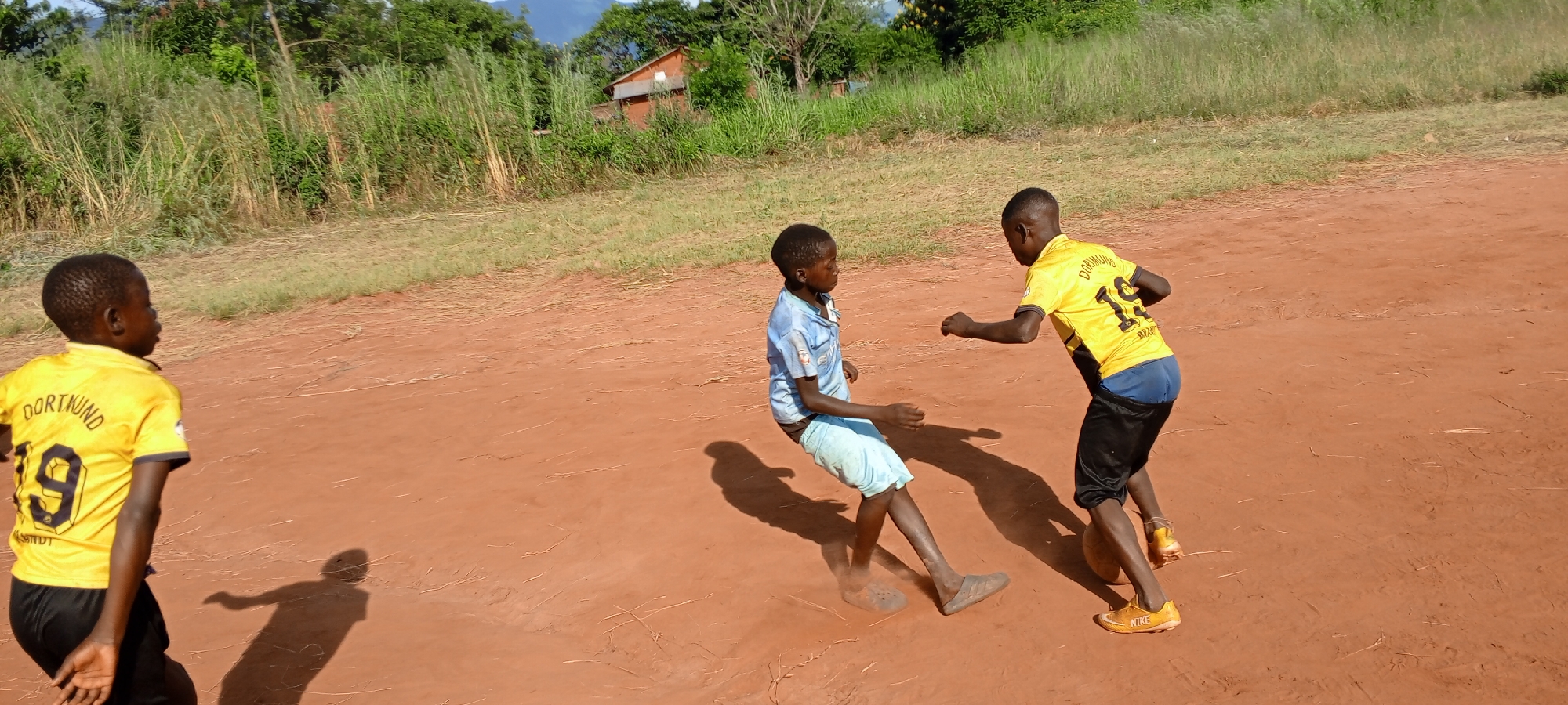 Three young boys are on a dirt field. The boy on the left is wearing a yellow shirt with "Dortmund 19" on the back, and black shorts. The boy in the center is wearing a blue shirt and light blue shorts. The boy on the right is wearing a yellow "Dortmund 19" shirt, blue shorts and orange shoes, and is kicking a soccer ball. There is grass and a few buildings visible in the background. The location is Ngalula, Democratic Republic of the Congo.