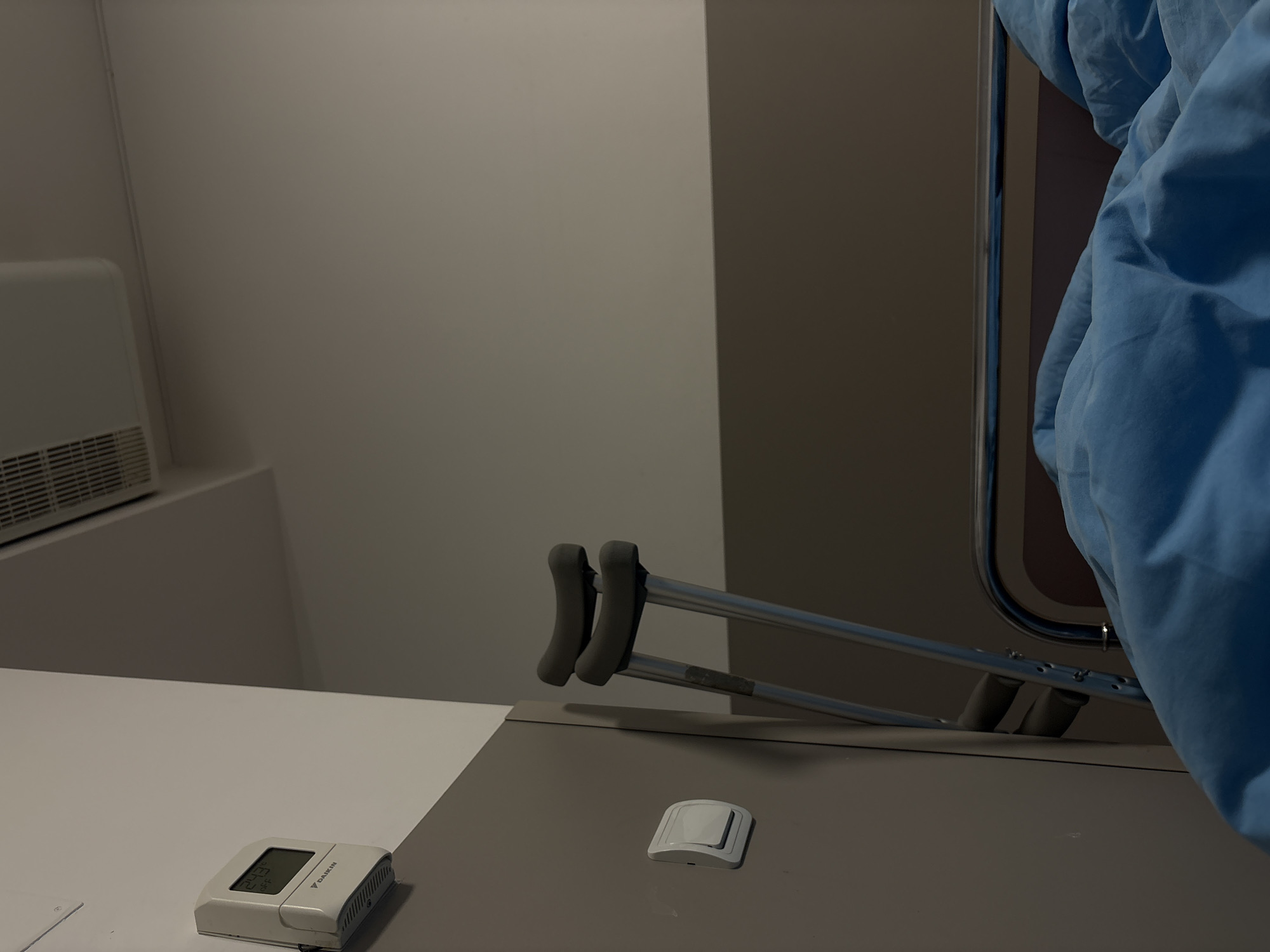 The image depicts an indoor scene with a focus on medical or healthcare elements. In the foreground, there is a white countertop with a digital thermostat and a small, square white object that appears to be a light switch or sensor. Behind the counter, a pair of crutches leans against a light gray wall or door, partially obscured by blue fabric, which suggests a curtain or medical gown. The environment is sterile, and the room appears to be in a hospital or clinic. The location in the city of Sofia, Bulgaria is not discernible from the image.