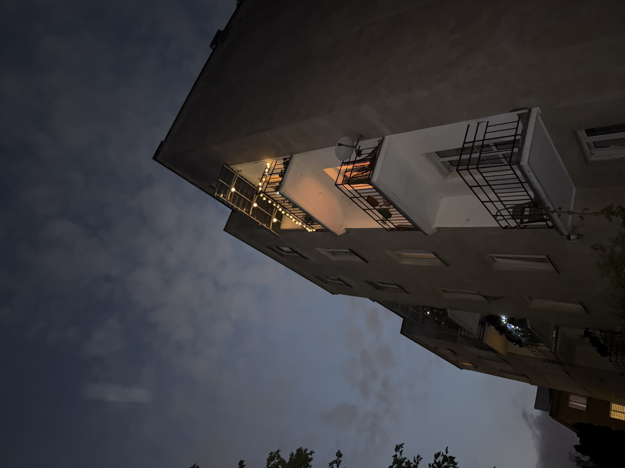 A low-angle, wide shot captures the facade of a multi-story apartment building at dusk. The building's exterior is a muted gray, with several white balconies jutting out. The balconies are adorned with black metal railings. One balcony, on the upper floors, is decorated with a string of warm-toned fairy lights. Another balcony below it has a satellite dish mounted on the railing. The sky above is a deep, twilight blue, with scattered, wispy clouds. Some foliage is visible in the lower left and right corners of the image. The lighting suggests it is either early morning or late evening.