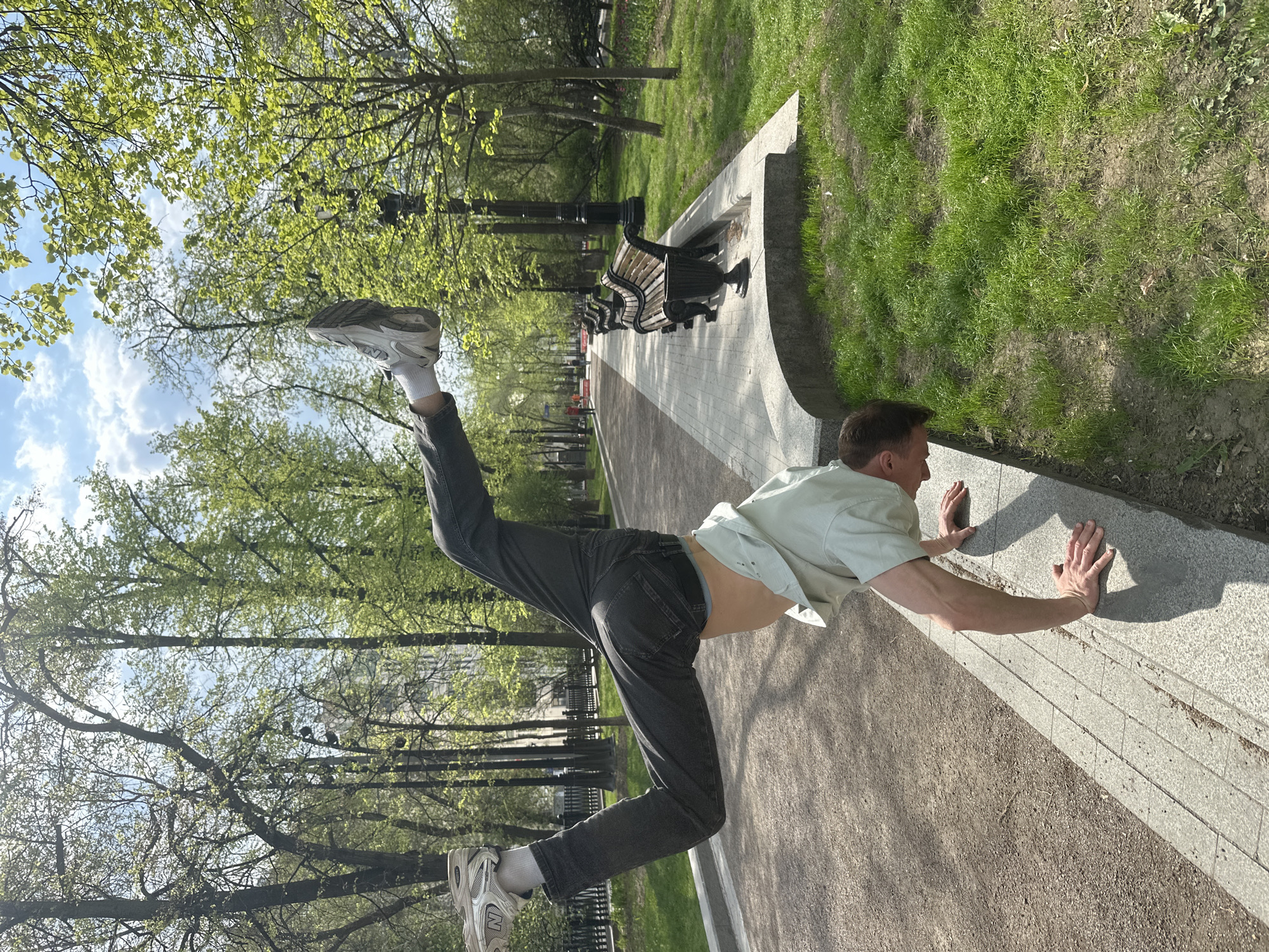A man wearing a light green short-sleeved shirt, dark jeans, white socks, and grey athletic shoes is performing a gymnastic-style pose in what appears to be a park. His hands are pressed against a grey concrete ledge, while his body is nearly horizontal, with one leg extended straight up and the other bent at the knee and pointed towards the sky. The scene is outdoors, likely in a park, with trees, grass, benches, and a paved pathway visible. The location is Moscow, Russia, as evidenced by the style of the park benches and street lamps. The weather appears to be sunny and pleasant.