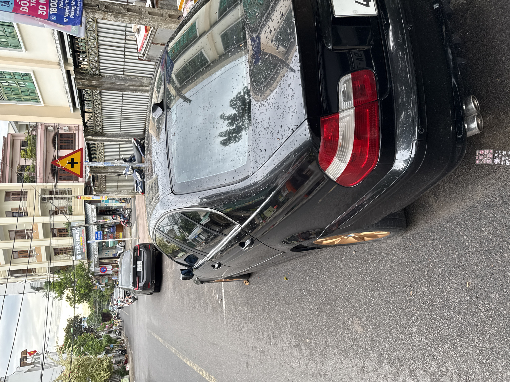 The image depicts a street scene in Nha Trang, Vietnam. In the foreground, a black sedan, possibly a BMW, is parked on the right side of the road. Raindrops are visible on the car's surface, indicating recent precipitation. The car has a license plate that is partially visible. Further down the street, another black sedan is visible. Several motorcycles are also present, with some riders on them, suggesting ongoing traffic. Buildings line both sides of the street, typical of an urban environment. One building prominently displays signage for "TUẤN NGUYỄN" with an Apple logo, indicating a business. A yellow triangular road sign with a black cross symbol is posted on a pole on the left side of the street, warning of an intersection. The sky is overcast, contributing to the muted lighting of the scene. The road appears to be asphalt.
