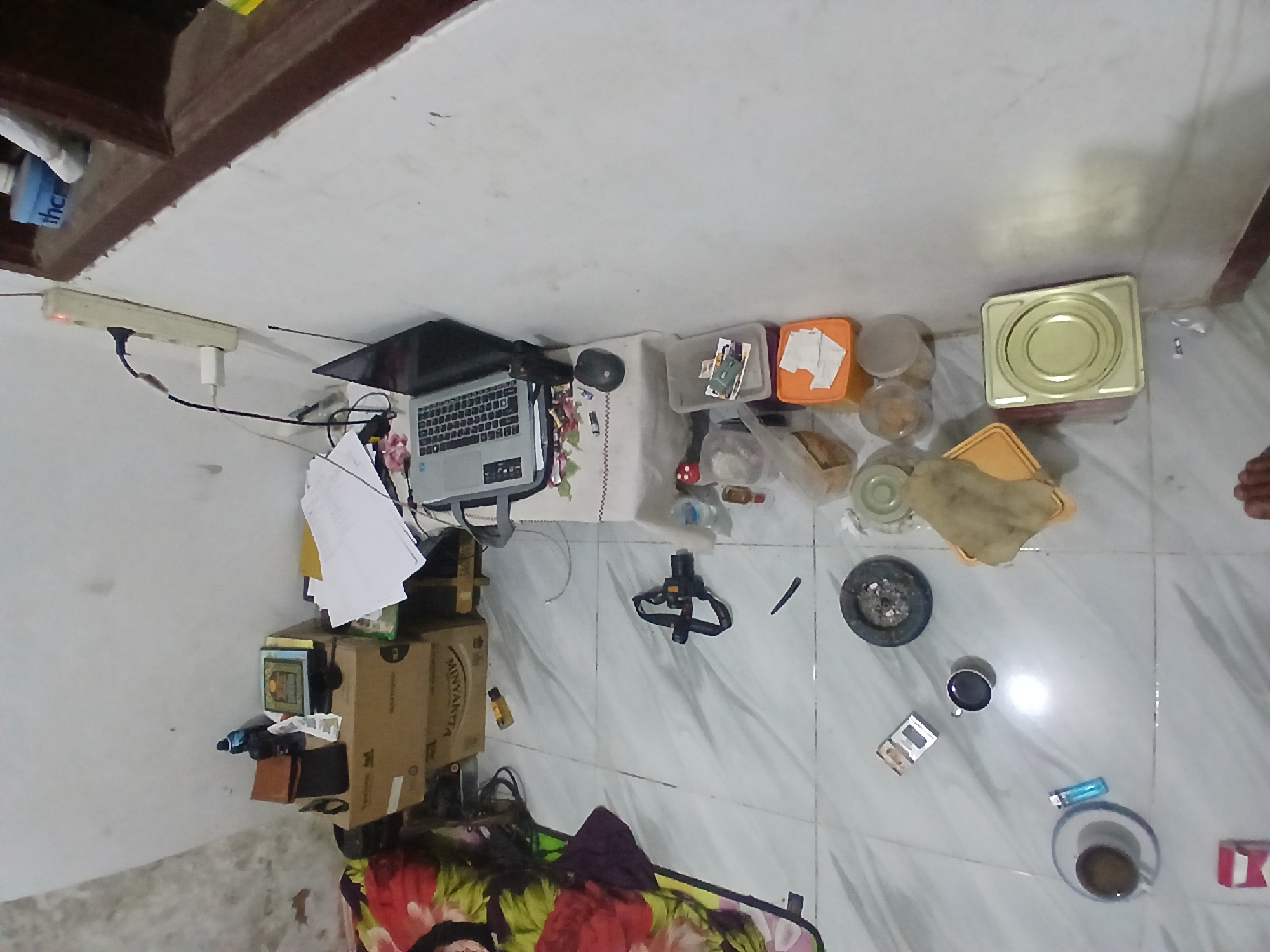 The image depicts a cluttered indoor scene, possibly a room in Kademangan Kulon, Indonesia, characterized by a white tiled floor and white walls. On the floor, a blanket with a floral pattern is visible on the left. Scattered items include a black camera with a strap, an ashtray filled with cigarette butts, a cigarette pack, a lighter, and two cups, one containing a dark liquid. A small, light-colored cigarette box is also present.
To the right, a table is set up with a laptop, a computer mouse, and various containers, including a grey plastic bin and an orange plastic container with white paper items. Several other plastic containers, some transparent and some opaque, are also present, containing small items. A yellow fabric is draped over a yellow plastic tray. Further to the right, a light-colored metal container with a circular indentation sits on the floor. Electrical outlets and cords are visible on the wall. A dark wooden window frame with a brown ledge is situated in the upper right corner of the image.