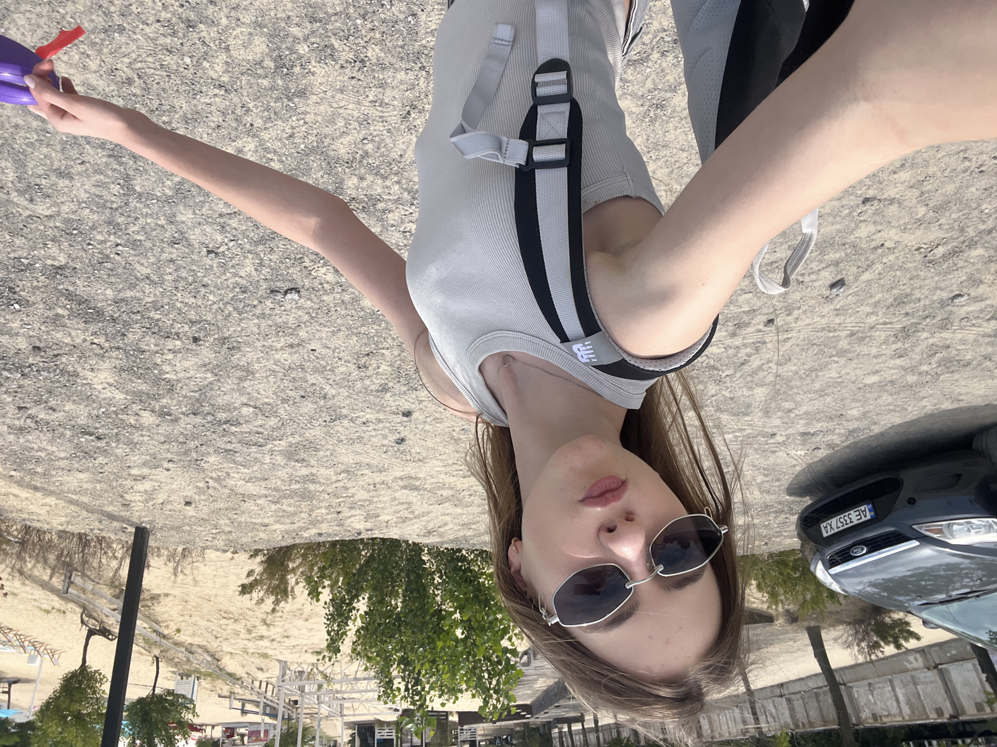 A young woman with long, brown hair and sunglasses is taking a selfie in an outdoor, sandy environment. She is wearing a light gray tank top and a backpack with black and gray straps. To her left, a dark gray Ford Focus car with license plate "AE 3357 XA" is parked on a gravel surface. In the background, there is a sandy beach area with wooden structures, trees, and lounge chairs. The overall scene appears to be a recreational area near a body of water. The specific location in the city of Yelyzavetivka, Ukraine is not identifiable from the image alone.