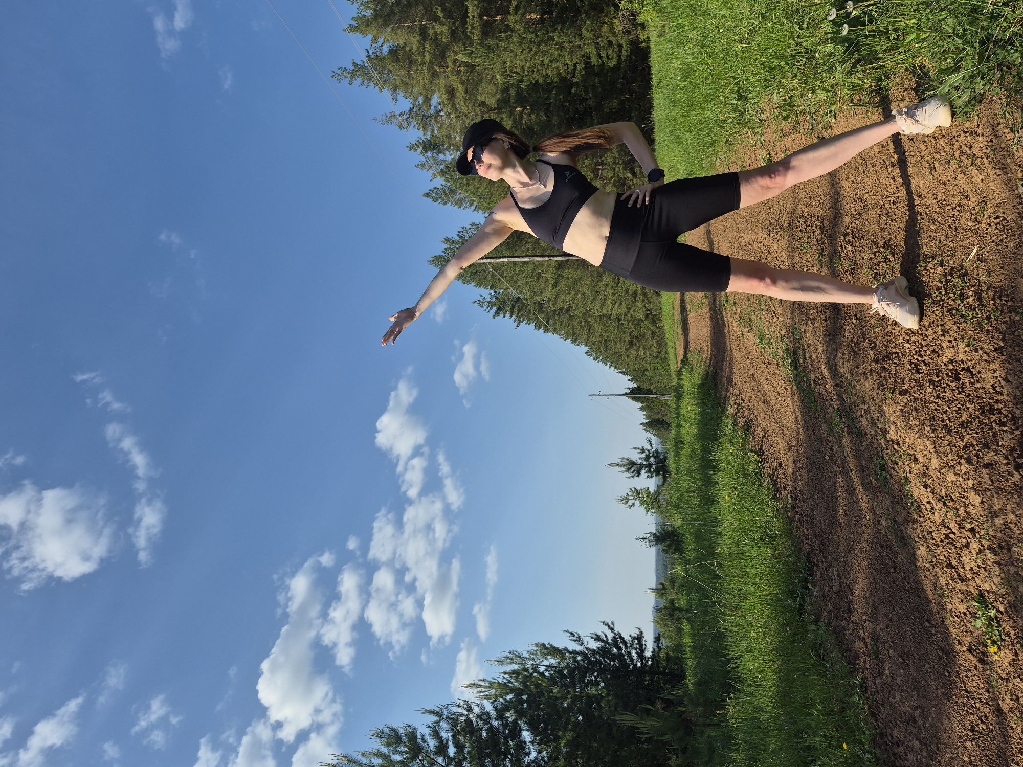 A woman in a black sports bra and black shorts, wearing a black cap and sunglasses, is standing on a dirt path. Her arms are extended, one towards the sky and the other at her hip. She is also wearing a wristwatch. The path is bordered by green grass and trees. The sky is blue with white clouds. The location appears to be a rural or wooded area, not in the city.