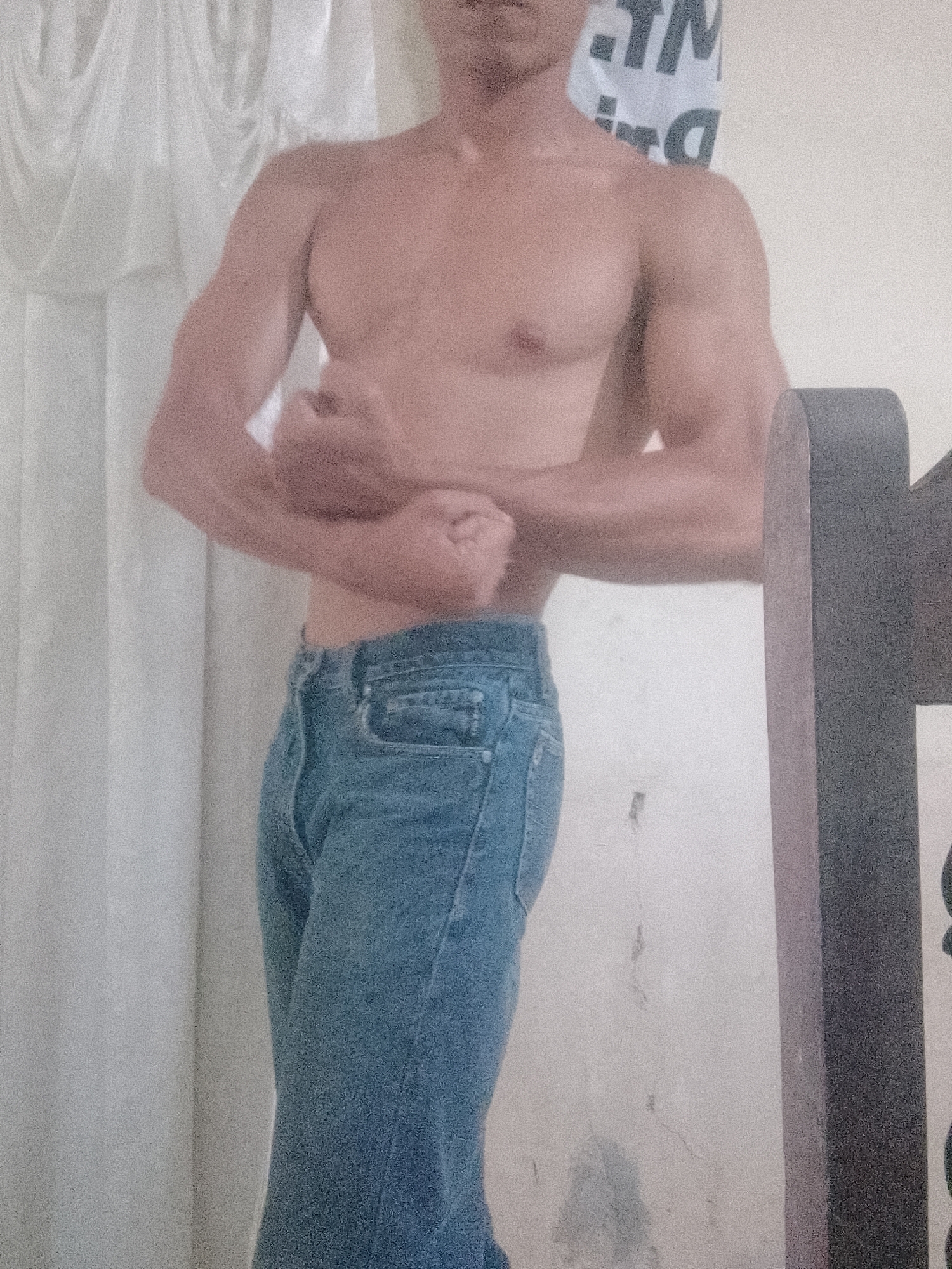 A shirtless man wearing jeans is standing indoors. His arms are bent and his fists are clenched in front of him. He is positioned in front of a white wall with a white curtain to his left. To his right, there is part of a dark gray object, possibly furniture. The backdrop behind the man includes a poster or sign with text on it. The exact location in Juan Griego, Venezuela cannot be determined from this image.