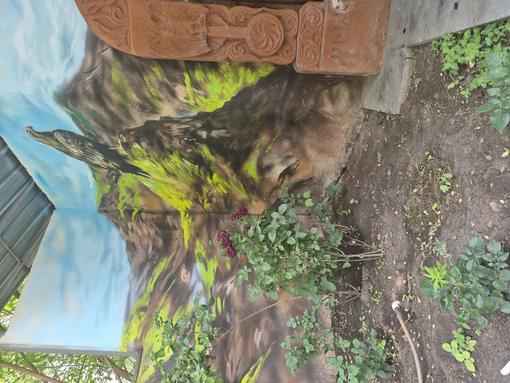 The image shows a corner of an outdoor space with a painted mural on the walls and a carved stone monument. The mural depicts a mountainous landscape with a large bird of prey perched on a rock, rendered in an airbrushed style with vibrant green foliage against brown rocks and a blue sky. To the right of the mural, a terracotta-colored stone monument with intricate carvings, including a circular floral pattern and what appears to be an inscription in Armenian script, stands next to the wall. In the foreground, several rose bushes with dark red blooms and lush green leaves are planted in the soil. A hose is visible lying on the ground. The area appears to be an outdoor garden or courtyard, potentially in Yerevan, Armenia, given the style of the monument and the script. The overall impression is of a decorative outdoor area with a cultural or artistic element.