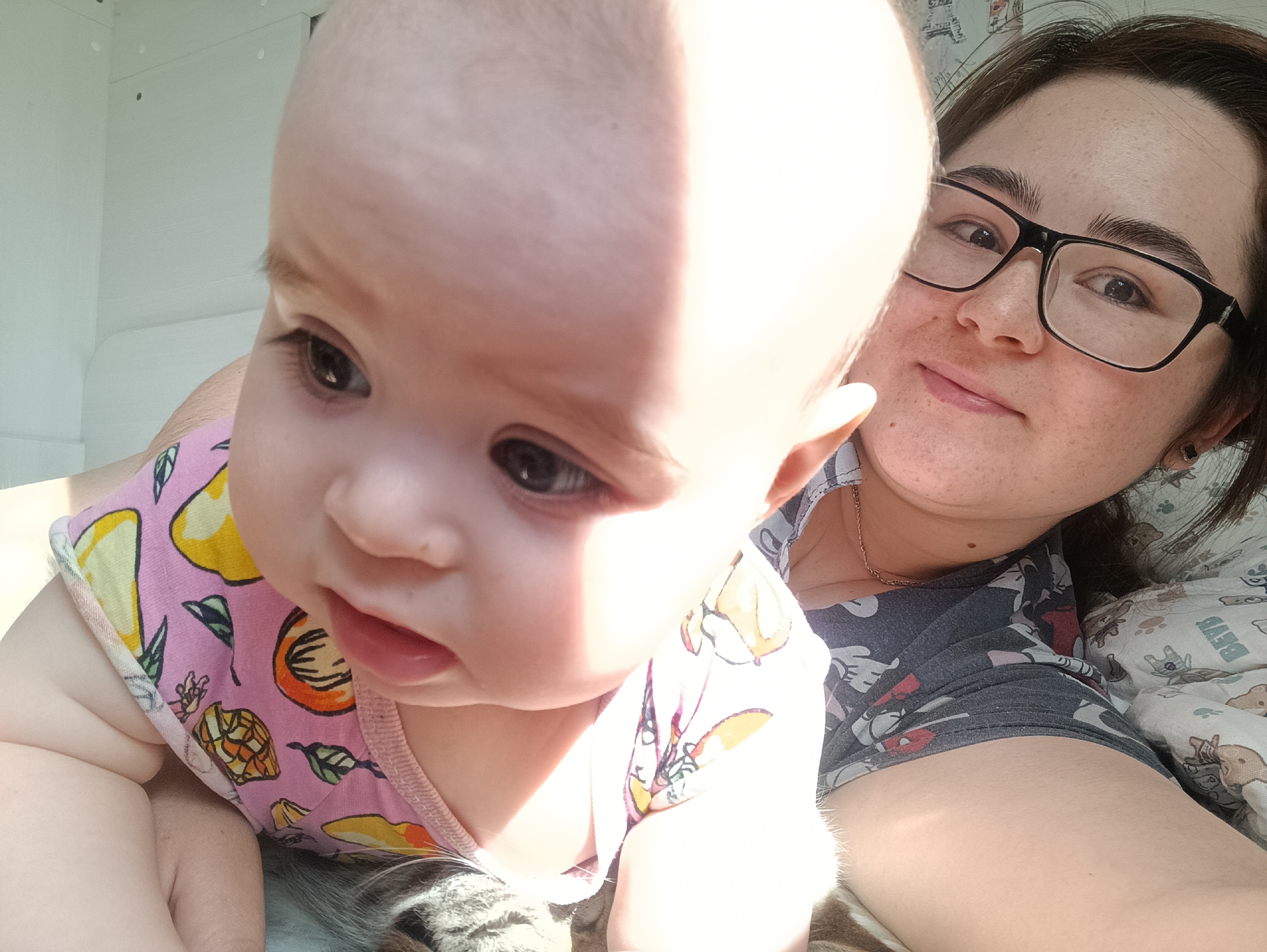 A close-up shot depicts a fair-skinned baby, wearing a pink onesie patterned with fruits, in the foreground. The baby is looking downwards and slightly to the left. Behind the baby, a light-skinned woman with dark hair and glasses is smiling at the camera. They appear to be lying on a bed with a patterned sheet. The location of Noviy, Russia, cannot be confirmed from the provided visual information.