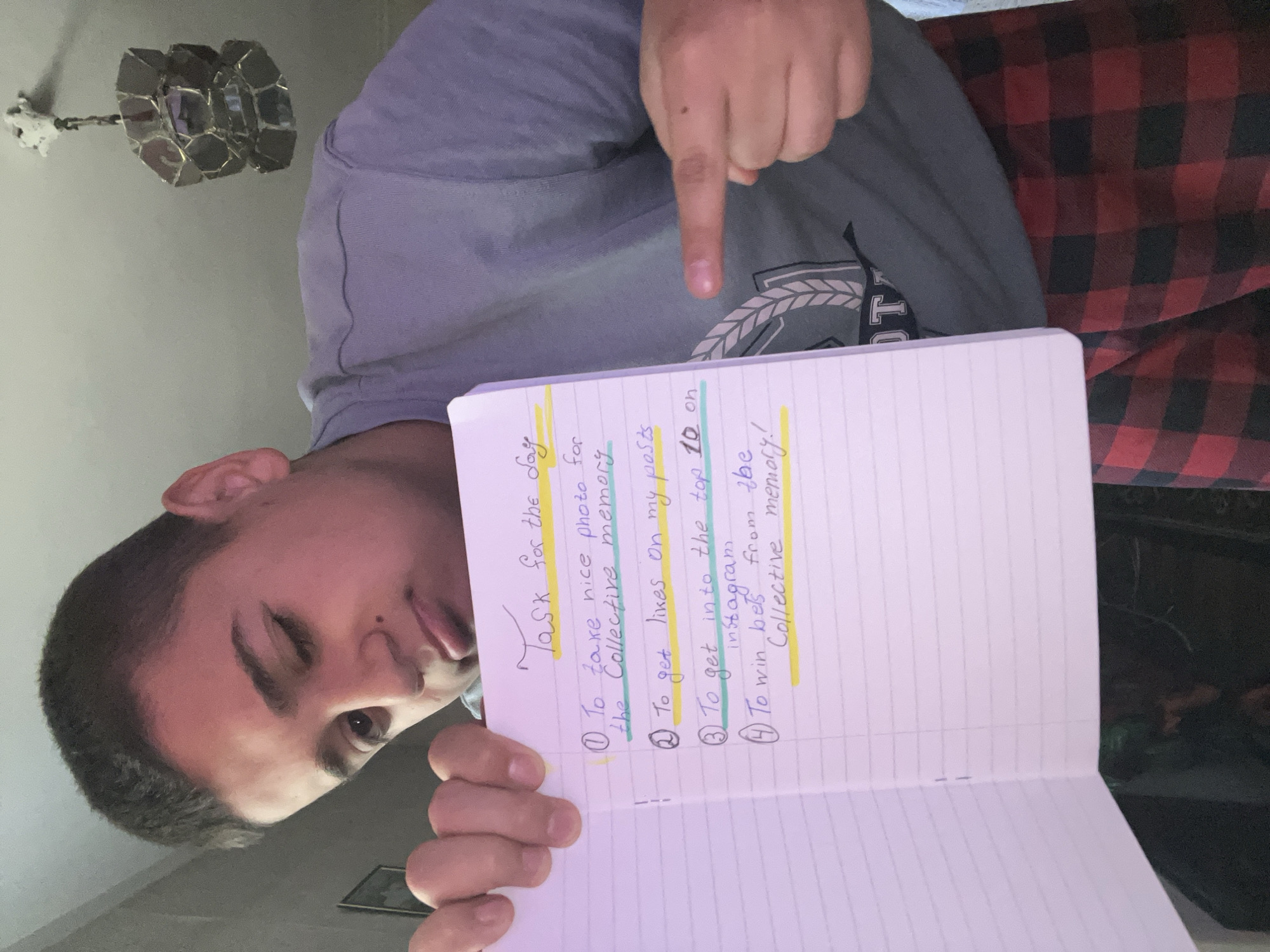 In an indoor setting in Lozarevo, Bulgaria, a young man is captured in a close-up, rotated image. He has dark hair and is wearing a grey t-shirt over what appears to be a red and black plaid fabric, possibly a blanket or another garment. He holds an open, lined notebook in his left hand, pointing to its contents with his right index finger. His expression suggests amusement or playfulness, featuring a slight smirk and a wink from his left eye.
The notebook displays a handwritten list titled "Task for the day," with the title highlighted in yellow. The numbered tasks are:
1. "To take nice photo for the Collective memory" (highlighted in yellow).
2. "To get likes on my posts." (highlighted in yellow).
3. "To get into the top 10 on Instagram" (highlighted in blue).
4. "To win bets from the Collective memory!" (highlighted in yellow).
In the background, a light-colored wall is visible, with a metallic, geometric light fixture attached to the ceiling. The lighting suggests an indoor setting, likely during the day or under artificial light.