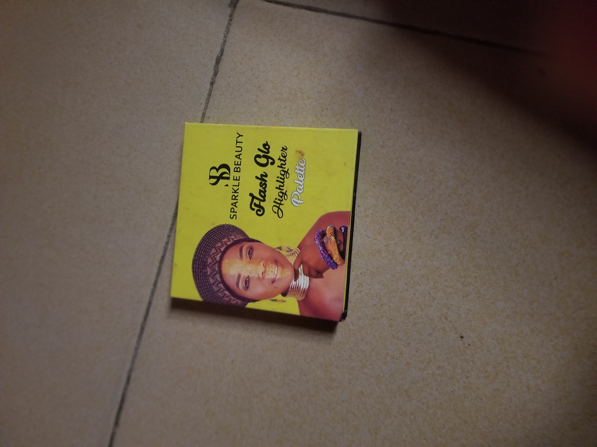This media file shows a close-up of a yellow makeup palette on a tiled surface. The palette features a woman's face, adorned with traditional jewelry and headwear, a common representation of beauty and culture in Nigeria. The text on the palette reads "SPARKLE BEAUTY," "Flash Glo Highlighter Palette." The lighting suggests it's daytime, with no specific weather details discernible. The setting appears to be an indoor location, given the tiled floor, possibly a home or a shop in Jauro Yinu, Nigeria. The woman's expression is one of a cheerful smile, conveying a sense of joy and confidence. The overall impression is one of vibrant and culturally inspired beauty products.
