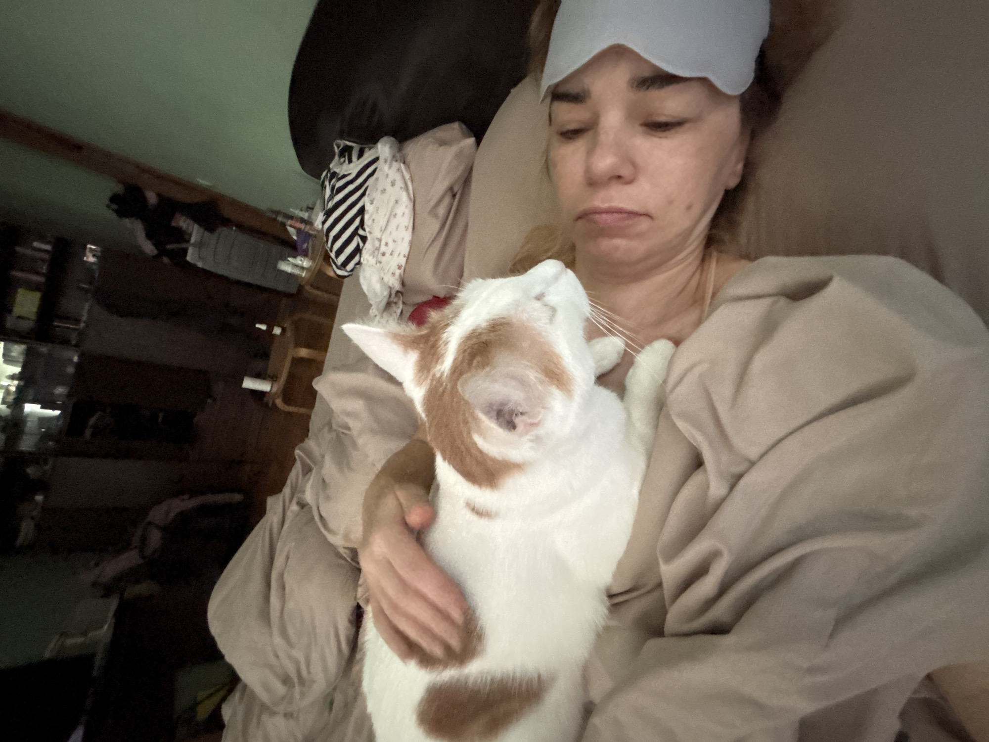 The image depicts a close-up, high-angle shot of a person and a cat in bed. The person is lying on their back, partially obscured by blankets and a light blue eye mask. Their eyes are closed, suggesting they are asleep or resting. A white and orange cat is positioned on the person's chest, its head turned towards the person's face, with its eyes closed. One of the person's hands is gently resting on the cat's back. The bedding is a neutral beige color. In the background, dimly lit shelves with various items are visible, along with a dark wooden floor. The scene appears to be an indoor bedroom setting.