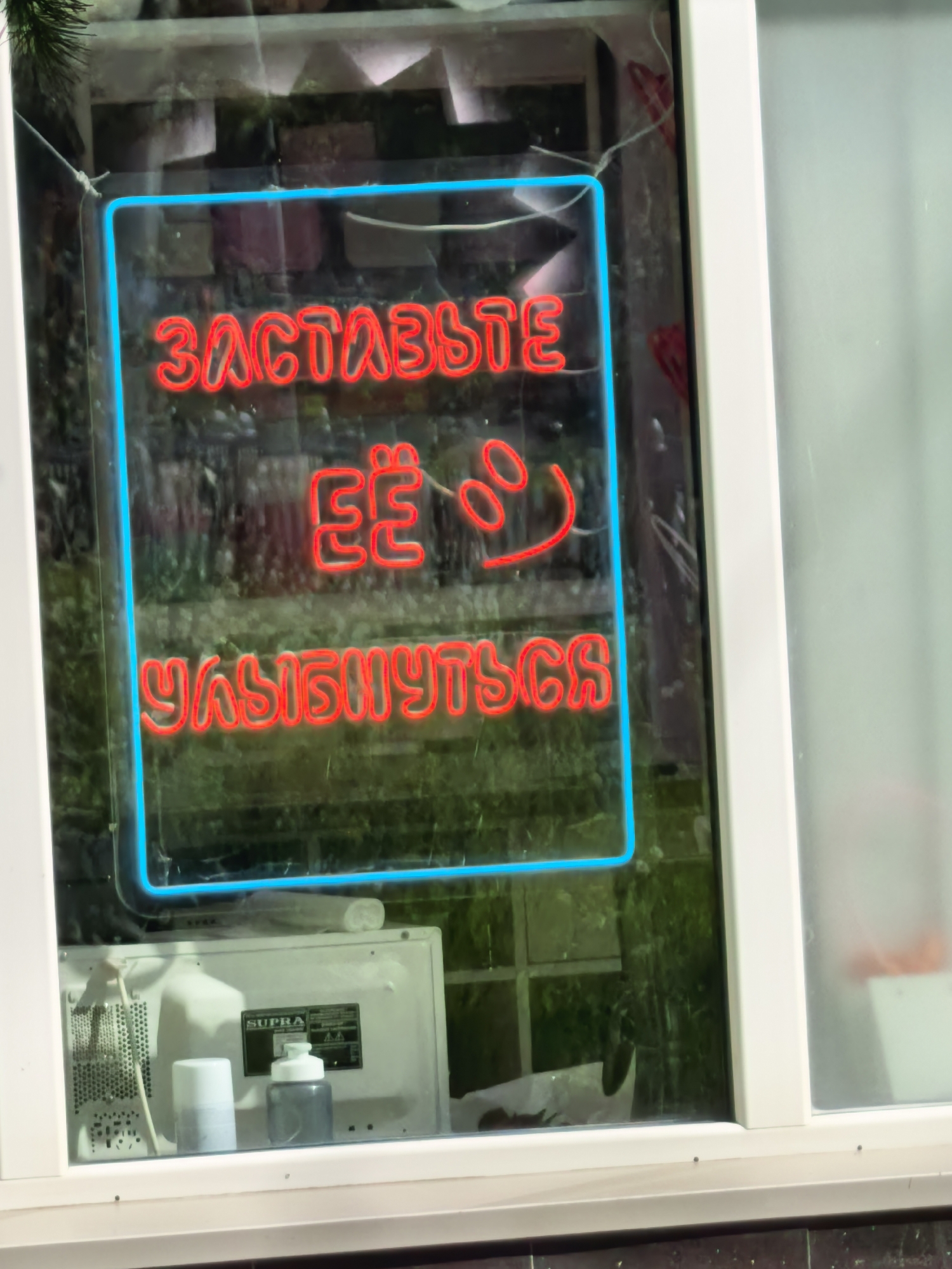 A daytime shot captures a window in Sterlitamak, Russia, displaying a neon sign with a blue frame. The red neon text within reads "ЗАСТАВЬТЕ ЕЁ" followed by a smiley face and "УЛЫБНУТЬСЯ" in Cyrillic, translating to "Make her smile" in English. Behind the sign, the blurred view through the window reveals brickwork, foliage, and an interior setting. Below the sign, a SUPRA-branded appliance is visible, along with two cylindrical containers, one white and the other dark gray. The window itself is framed in white.