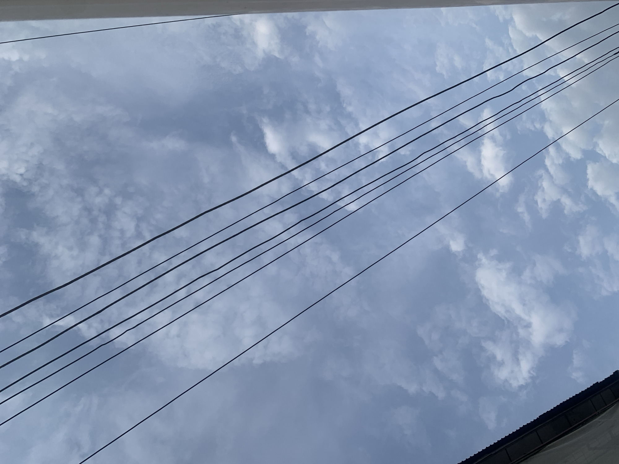 An upward-angle outdoor shot captures a sky filled with scattered, white, cumulus clouds against a pale blue backdrop. Several dark, thin electrical wires are diagonally strung across the frame, from the top left to the bottom right. In the lower left corner, the dark, corrugated metal roof of a building is visible, angled upwards. The right edge of the frame shows a sliver of a white, vertical structure, possibly a wall or pillar. The scene is set in Elelenwo, Nigeria.