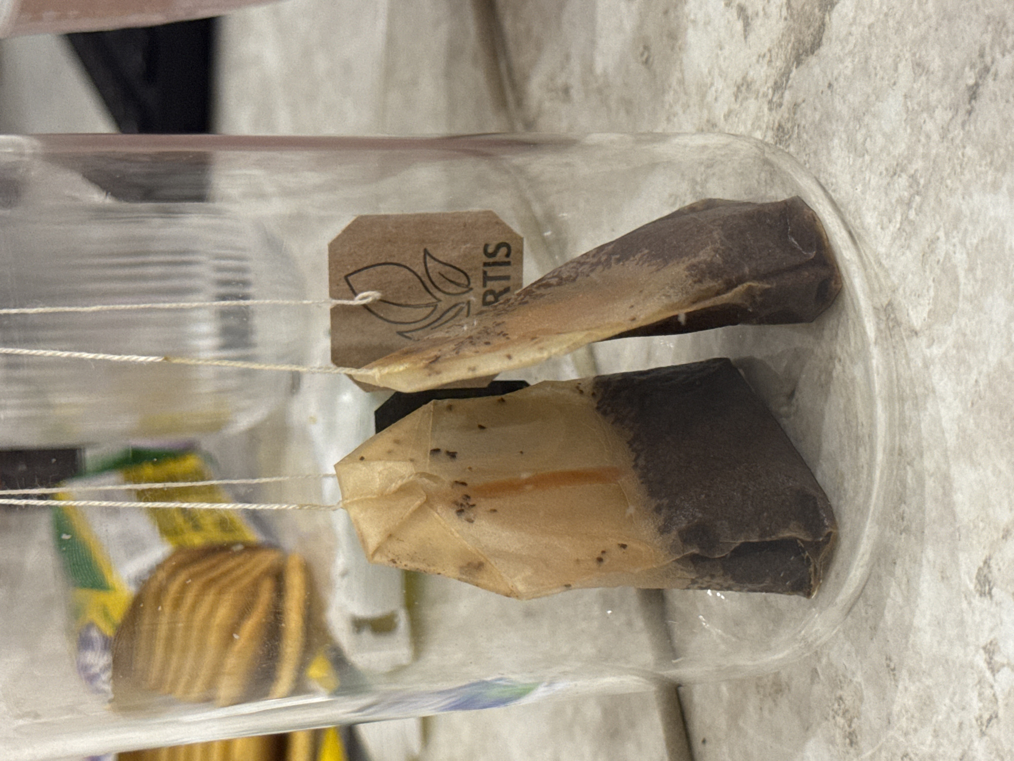 Two used tea bags are suspended by strings inside a clear glass container. The tea bags are stained dark brown at the bottom, indicating they have been steeped in liquid. The strings are white and appear to be made of cotton. A small brown tag is attached to one of the tea bags, displaying a logo and the text "RTIS". The glass container is placed on a speckled, light-colored surface, possibly a countertop. In the background, out of focus, are other objects, including what appears to be a package of cookies or crackers and a partially visible pink object. The scene is indoors, likely in a kitchen or similar domestic setting. There is no visible indication of the location in Saratov, Russia.