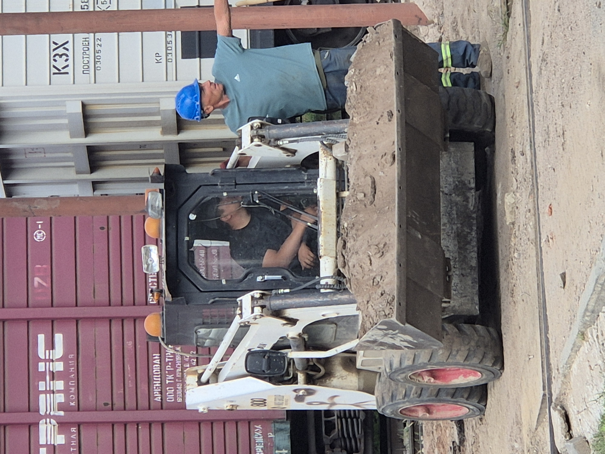 The image captures a construction scene, likely at a railway yard, depicting a Bobcat skid-steer loader filled with dirt. Two individuals are present. One, wearing a blue hard hat and a teal t-shirt, stands to the right of the loader, gesturing with his right arm. The other, partially visible inside the loader's cab, is wearing a black shirt and appears to be operating the machinery. The background reveals the side of a burgundy freight train car and a white container with Cyrillic lettering, including "K3X" and numbers. The ground is composed of dirt and gravel. The presence of Cyrillic text suggests a location in a Russian-speaking region, and while the provided context specifies Kokshetau, Kazakhstan, the image itself does not contain definitive visual cues to confirm this specific location.