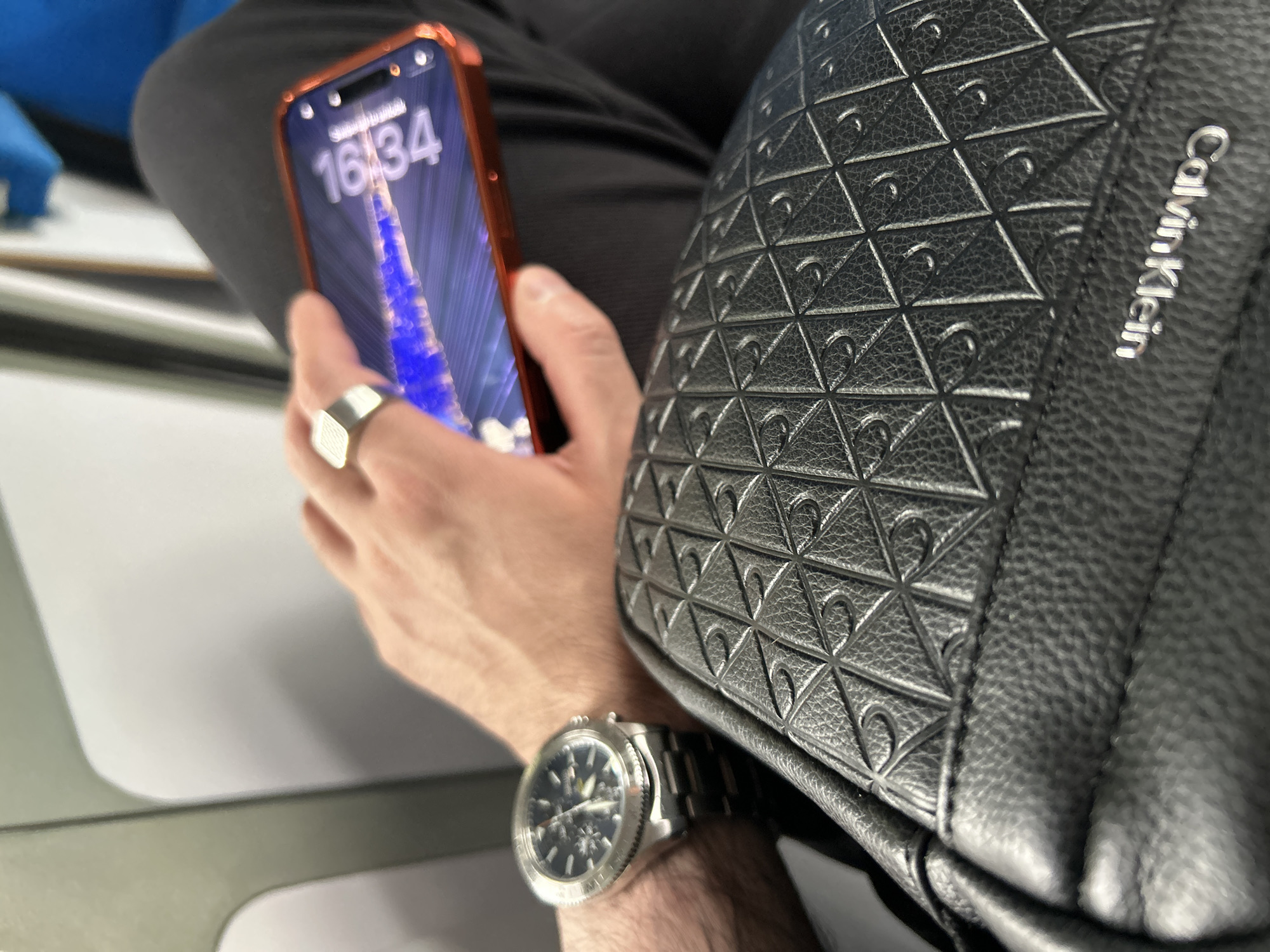 A person sits with a black Calvin Klein bag resting on their lap. They hold an orange-cased smartphone displaying the time "16:34" and an image of a blue light shooting up into the night sky. The person wears a silver signet ring on their left hand and a silver wristwatch on their left wrist. The scene takes place on a gray and white surface, likely inside a vehicle or building. The location is Briesen (Mark), Germany.