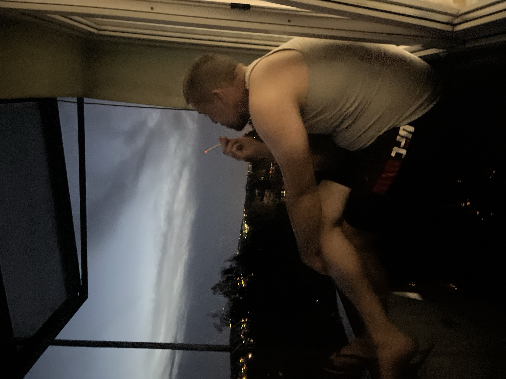 An adult male is seated outdoors at dusk, looking out over a city skyline. He is wearing a light-colored tank top and dark shorts. He is holding a cigarette to his mouth and appears to be smoking. The background shows a dark sky with some clouds and the lights of a city in the distance. The man is positioned on a balcony or porch, as indicated by a railing and some structural elements of a building. The overall lighting suggests it is late evening or early morning.