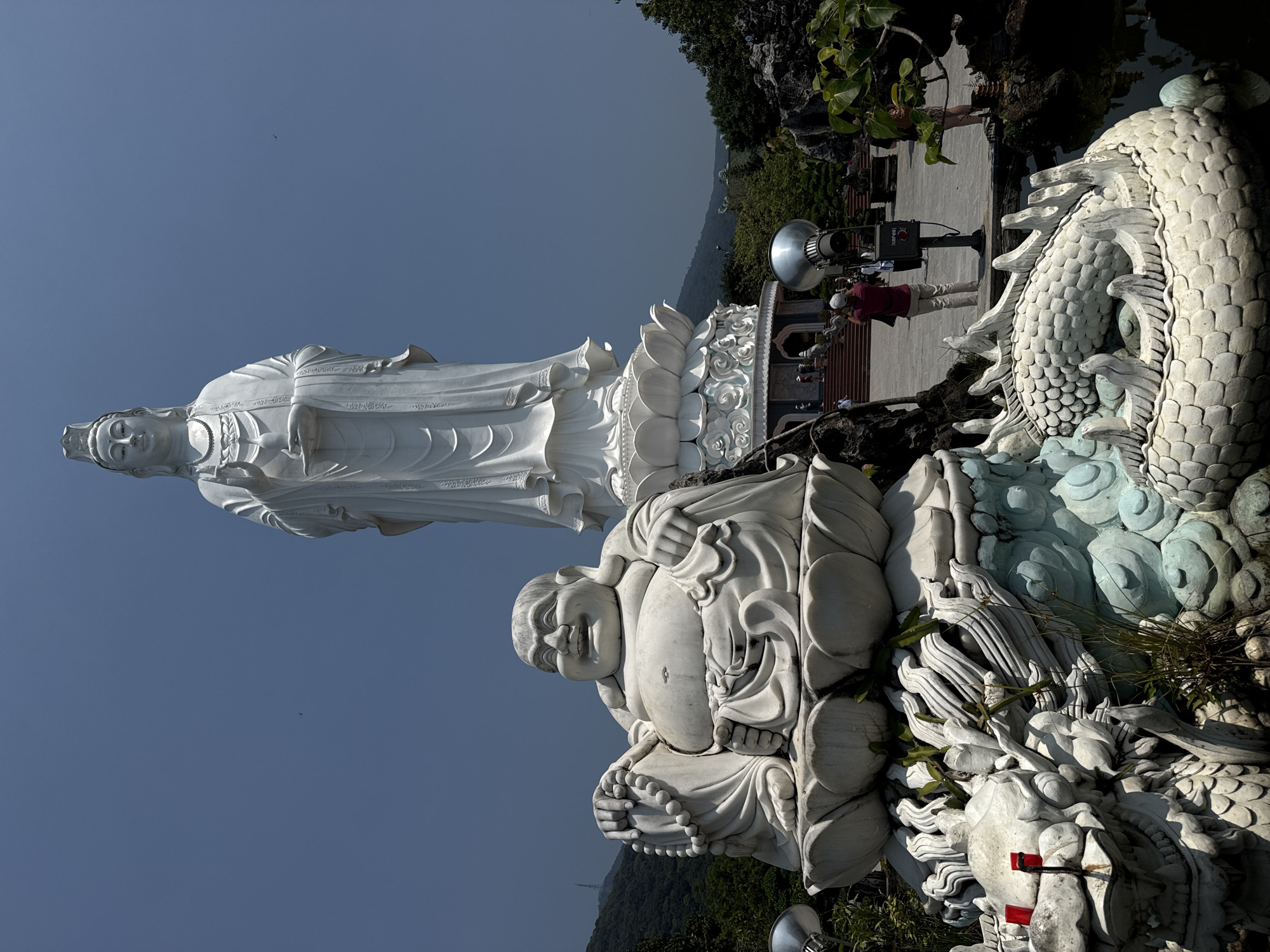 This image depicts a wide shot of the Linh Ung Pagoda on Son Tra Peninsula in Đà Nẵng, Vietnam, featuring prominent Buddhist statuary under a clear sky.
The primary subject is a colossal, white, standing statue of Guan Yin (Bodhisattva of Mercy), positioned in the midground and rising prominently against the light blue sky. She wears flowing robes, a necklace, and a crown, with her right hand raised and her left hand holding a small bottle. The statue rests on a multi-tiered lotus pedestal, which is part of a larger, elaborate white and light blue base structure with multiple steps leading up to it.
In the foreground, to the left, is a large, white, seated statue of the Laughing Buddha (Hotei). He is portly, depicted with a broad smile, holding a mala (prayer beads) in his left hand while his right hand rests on his knee. This statue is also situated on a lotus-shaped base.
Below the Laughing Buddha statue, intricately carved white and light blue dragon sculptures are visible, forming a decorative base or surrounding a dark water feature, possibly a pond, which extends into the lower right of the frame. One dragon's head in the lower left exhibits red accents on its snout.
Several individuals are present in the midground. One person, dressed in a red shirt, tan pants, and a white cap, is actively photographing the scene with a camera or phone, facing towards the Guan Yin statue. Other smaller figures are visible near the base of the Guan Yin statue, ascending the steps.
Two large, silver, conical lighting fixtures or speakers are positioned in the midground. One is to the left of the Laughing Buddha, and another, mounted on a grey pole with a small control box labeled "HARP," stands in front of the Guan Yin statue's base.
The ground consists of a paved stone courtyard leading towards the main statue's base. Green foliage, including various bushes, grasses, and tree leaves, surrounds the statues and the water feature, with some plant growth visible on the Laughing Buddha's base.
In the background, lush green mountains or hills rise under a clear, cloudless blue sky. A tall, slender structure, possibly a radio tower, is faintly visible on a distant mountain peak to the left. A small, dark, indistinct object, possibly a bird, is visible in the upper mid-left sky.