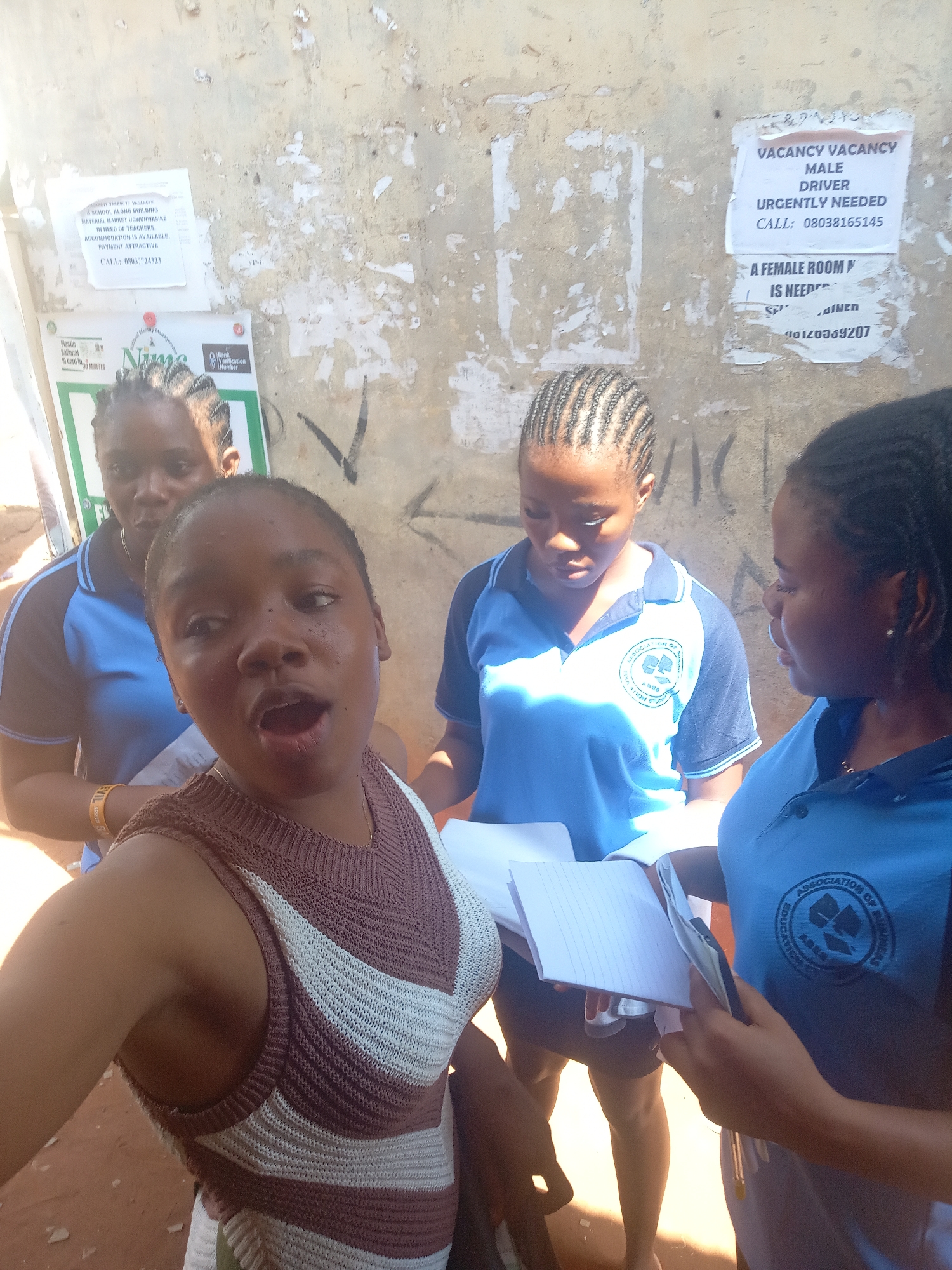 Four young women are captured in a bright, daytime outdoor setting in Nsugbe, Nigeria, with a weathered, light-colored wall serving as the backdrop. The woman in the immediate foreground, positioned to take a selfie, wears a brown and white patterned sleeveless knit top and has an open-mouthed, expressive look.
Behind her, three other young women are visible, all wearing matching light blue and dark blue polo shirts, indicative of school uniforms. The emblem on their shirts reads "ASSOCIATION OF BUSINESS EDUCATION STUDIES." The two women to the right are intently looking down at open notebooks and papers they hold, appearing engaged in a discussion or study session. One holds a pen, poised to write. The third woman, further to the left and slightly out of focus, also wears the uniform and is observing the scene.
The wall behind them is adorned with several tattered papers and posters, suggesting a public or frequently trafficked area. Visible text on these posters includes:
- "VACANCY VACANCY A SCHOOL ALONG BUILDING MATERIAL MARKET UGWUNWASIKE IN NEED OF TEACHERS, ACCOMMODATION IS AVAILABLE. PAYMENT ATTRACTIVE CALL: 08037724323"
- "Plastic National ID card in 30 minutes" alongside "NIMS" and "Bank Verification Number" logos.
- "VACANCY VACANCY MALE DRIVER URGENTLY NEEDED CALL: 08038165145"
- A partially obscured poster about "A FEMALE ROOM IS NEEDED" with a contact number starting "08120939207."
The overall scene suggests a casual gathering of students, possibly during a break or engaged in an outdoor learning activity, set against the backdrop of community notices.