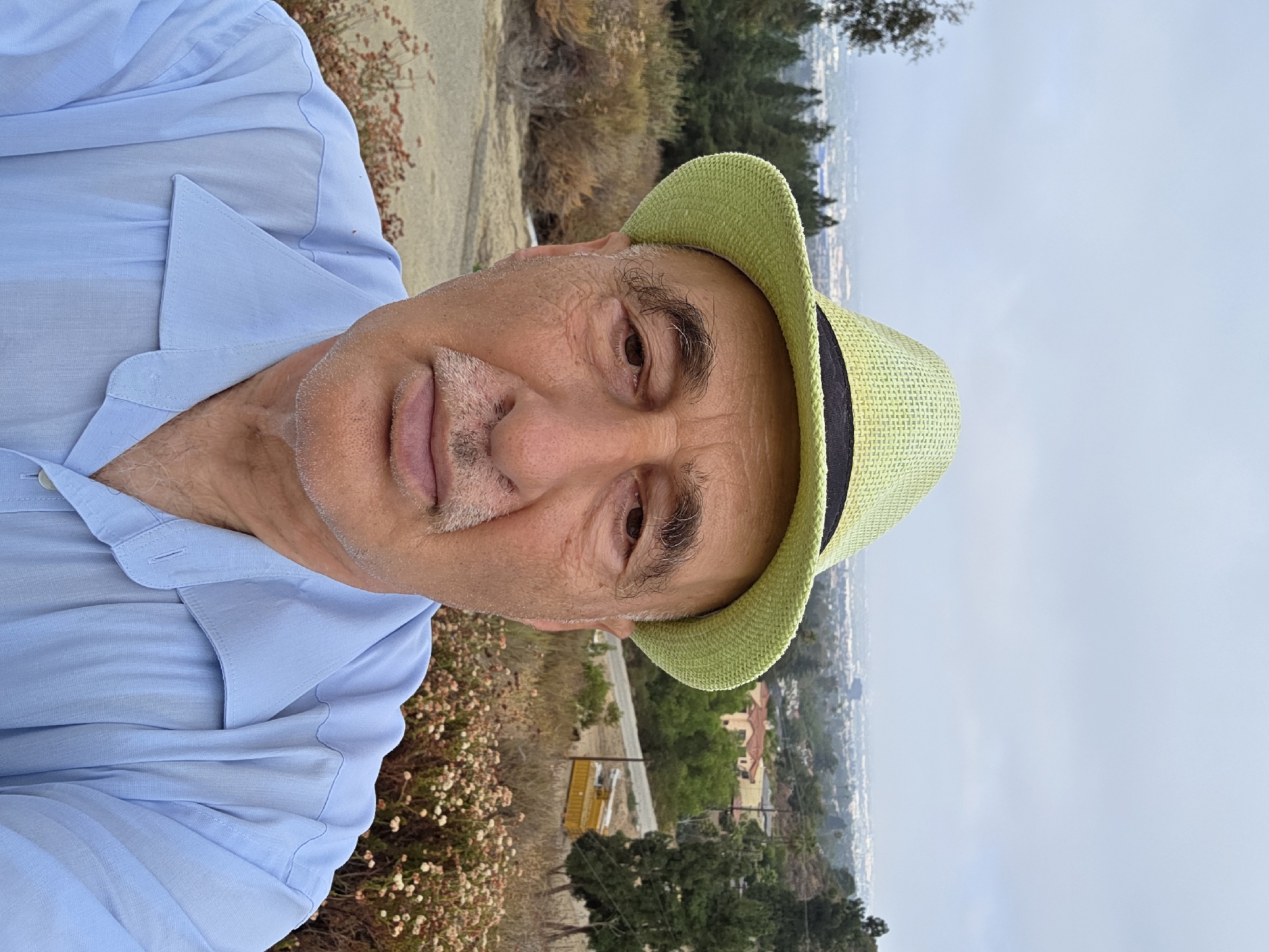 An elderly Caucasian man with a light skin tone is taking a selfie outdoors. He is wearing a light green fedora-style hat with a black band and a light blue collared shirt. The man has thinning gray hair, prominent eyebrows, and a mustache. His face is aged with visible wrinkles and some facial stubble.
The background appears to be a hillside overlooking a city, possibly Glendale, California, given the style of the houses and the general landscape which is characterized by dry brush, scattered trees, and a hazy, overcast sky. In the mid-ground, there is a road with a yellow vehicle, possibly a construction vehicle, parked on the side. The foreground is dominated by more dry brush and some small flowering plants. The overall impression is a suburban or semi-rural overlook within a metropolitan area.