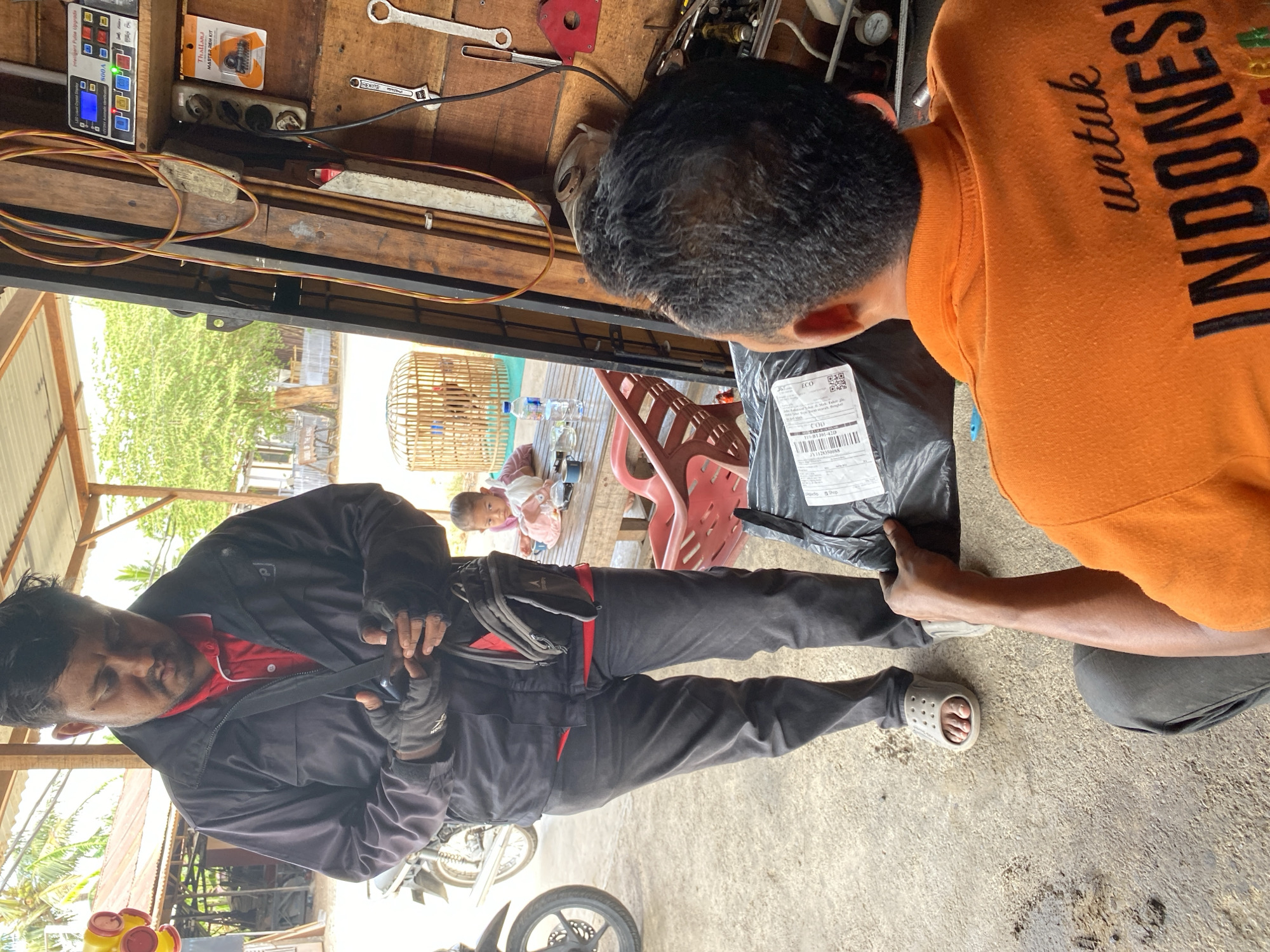 In Bayu, Indonesia, two men are engaged in a transaction in an outdoor, rustic setting. The man on the left, wearing a dark jacket over a red polo shirt and black pants, is holding a smartphone and appears to be interacting with a package. He has dark hair and a beard, and is wearing grey slip-on shoes. To his right, a child is seated at a wooden table, partially visible. Behind the child, a wooden bird cage contains a chicken. The man on the right, facing away from the camera, is wearing an orange polo shirt with the text "untuk INDONESIA" on the back. He is holding a black package, seemingly in the process of receiving it from the man on the left. The environment features wooden structures, a motorcycle on the left, and various tools and electrical equipment mounted on a wooden wall in the background. The ground is composed of concrete and dirt.