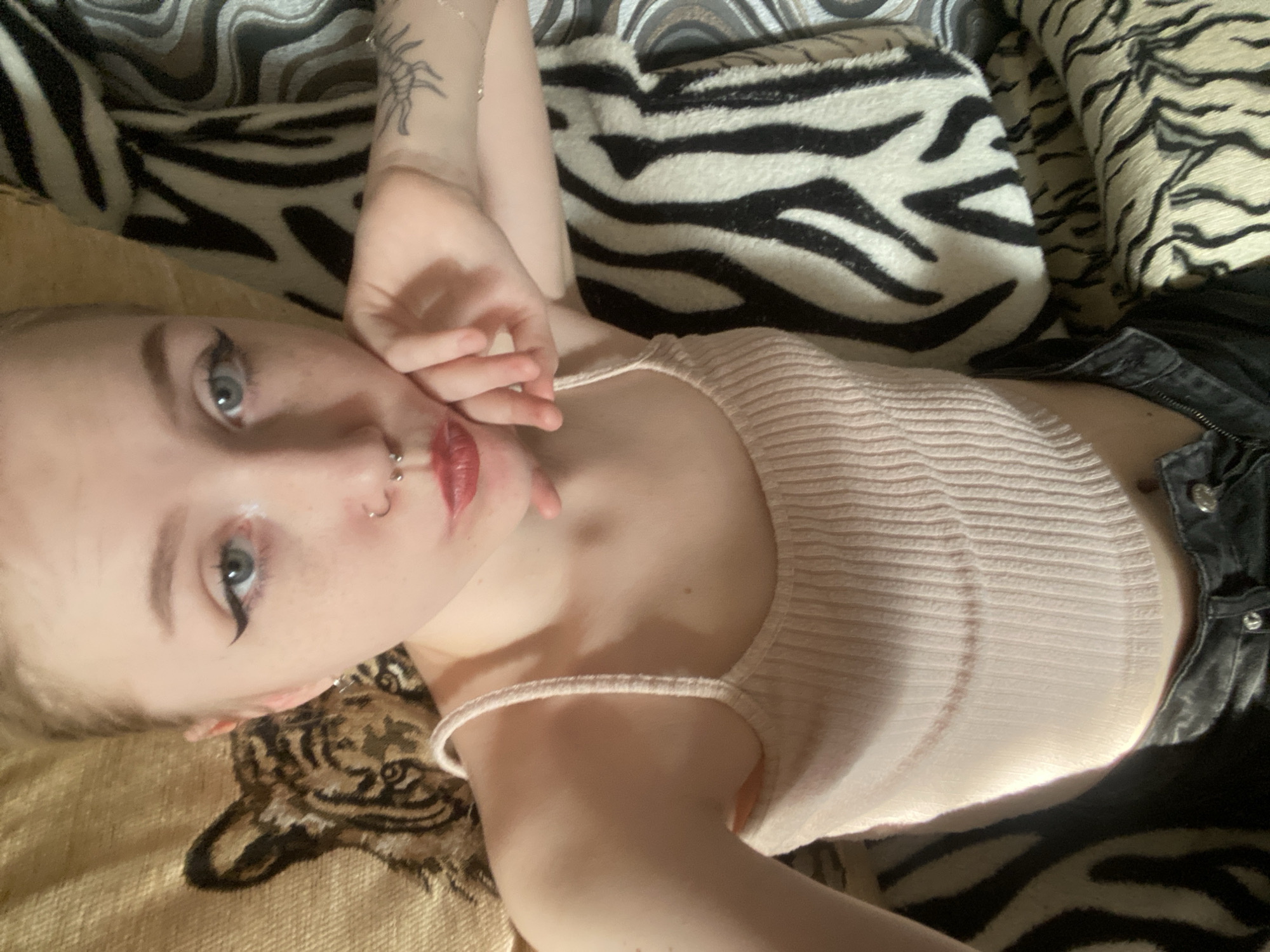 A young woman with fair skin, light blue eyes, and a nose piercing is lying on a patterned cushion. She is wearing a light pink ribbed tank top and dark jeans. Her left hand is raised near her face. The cushion appears to be a mix of zebra and tiger print patterns. The location is not discernible as Izhevsk, Russia.