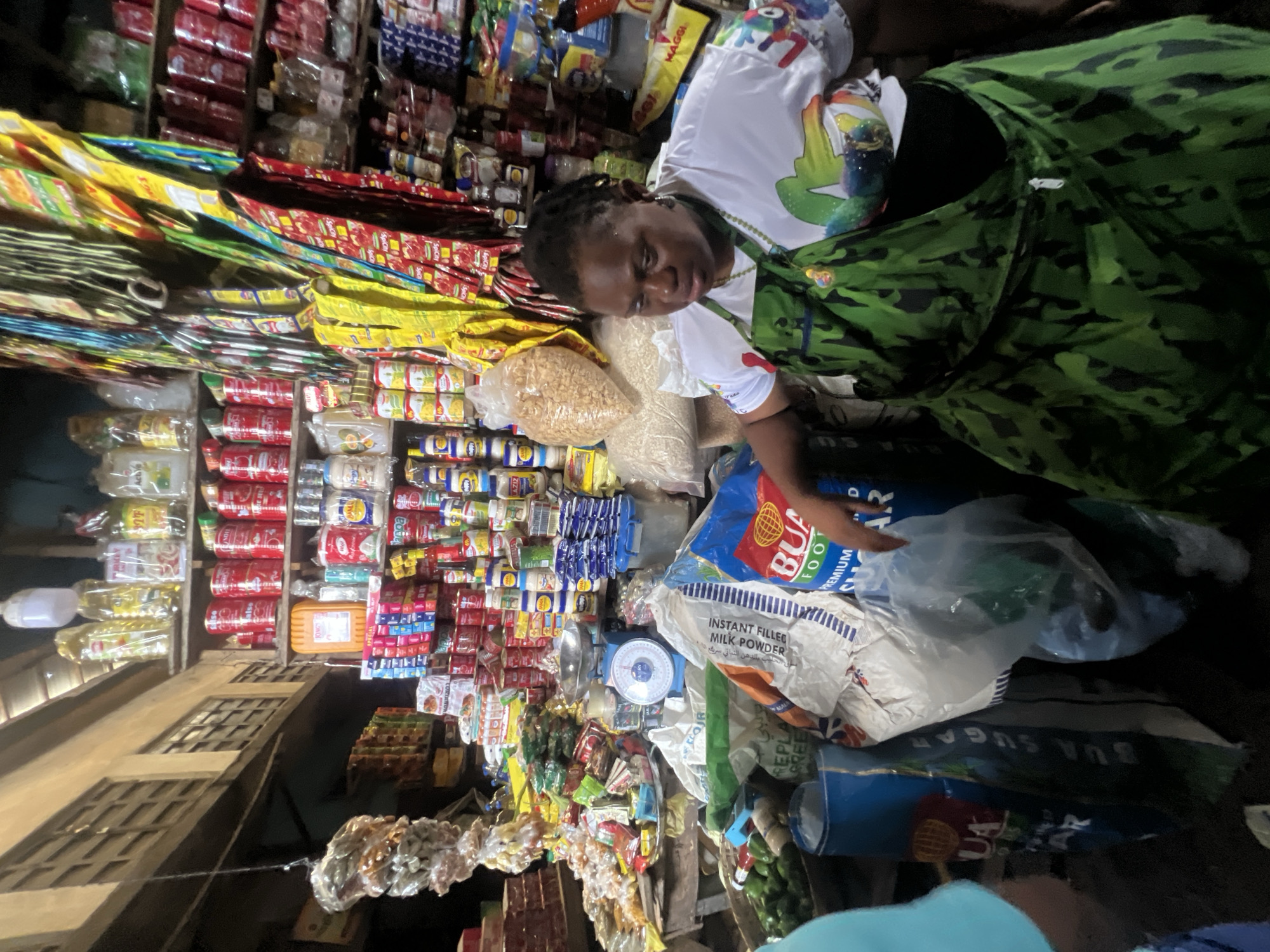 The image shows an individual, possibly a vendor, situated amidst a bustling Nigerian retail environment. The location appears to be a small shop or stall in Nkpor, Nigeria, evident from the various locally branded products prominently displayed. These products include bags of BUA sugar and milk powder, as well as an assortment of packaged foods and household goods. The individual seems to be tending to their merchandise, potentially weighing or preparing goods for a customer. The setting is densely packed with merchandise, suggesting a vibrant but space-constrained commercial area. The photograph depicts a scene of everyday commerce within Nkpor.