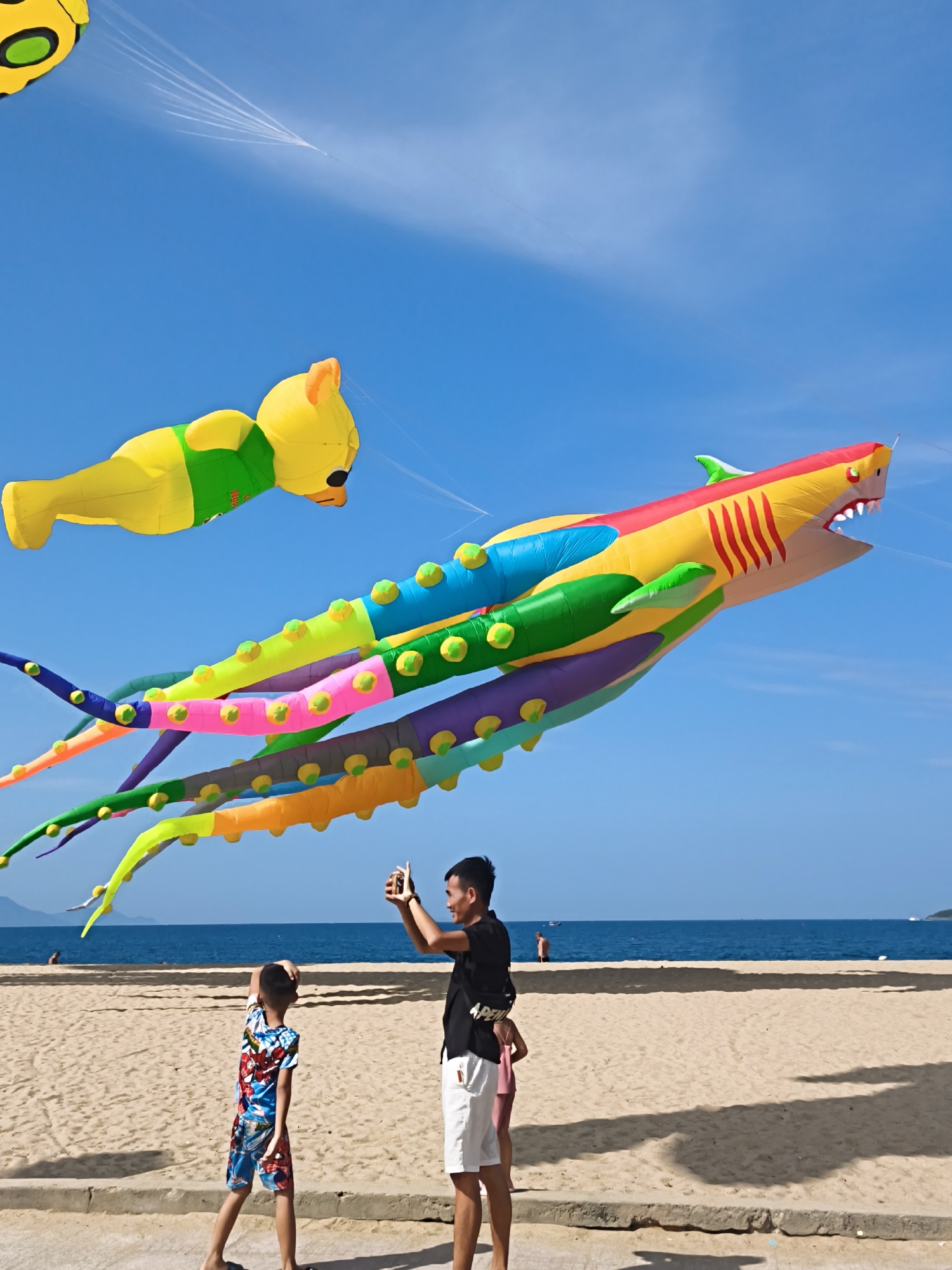 On a sunny day at a sandy beach in Nha Trang, Vietnam, a man and two children are enjoying the scenery. The man, wearing a black t-shirt and white shorts, is taking a photo with his phone of the vibrant, inflatable kites flying in the clear blue sky. One kite is shaped like a yellow bear, and another is a colorful shark with multiple long, segmented tentacles. The children, one in a patterned shirt and shorts, and another partially visible in a pink dress, are also looking up at the kites. The ocean stretches out behind them, with a few distant boats visible on the horizon. The sand is a light tan color, and a concrete walkway runs along the edge of the beach, where the people are standing. The scene is typical of a leisurely day at the beach, with the focus on the playful spectacle of the kites.