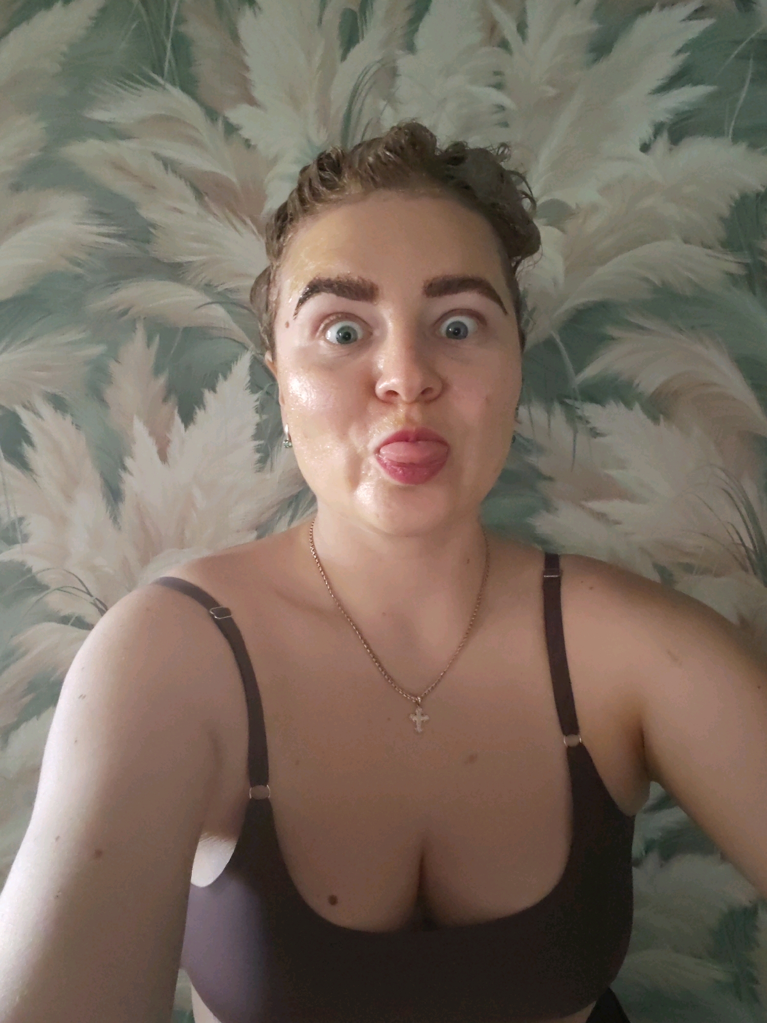 A woman with light skin and brown hair pulled back into curls, with a face mask on, makes a funny face with her tongue sticking out at the camera. She is wearing a dark bra with a thin chain necklace with a cross pendant. The background is a wallpaper with a pattern of feathery reeds in light green and cream colors. The woman's eyes are wide and her eyebrows are raised. The image appears to be taken indoors, possibly in a bathroom or bedroom. There is no information available regarding the location in the city of Izhevsk, Russia.
