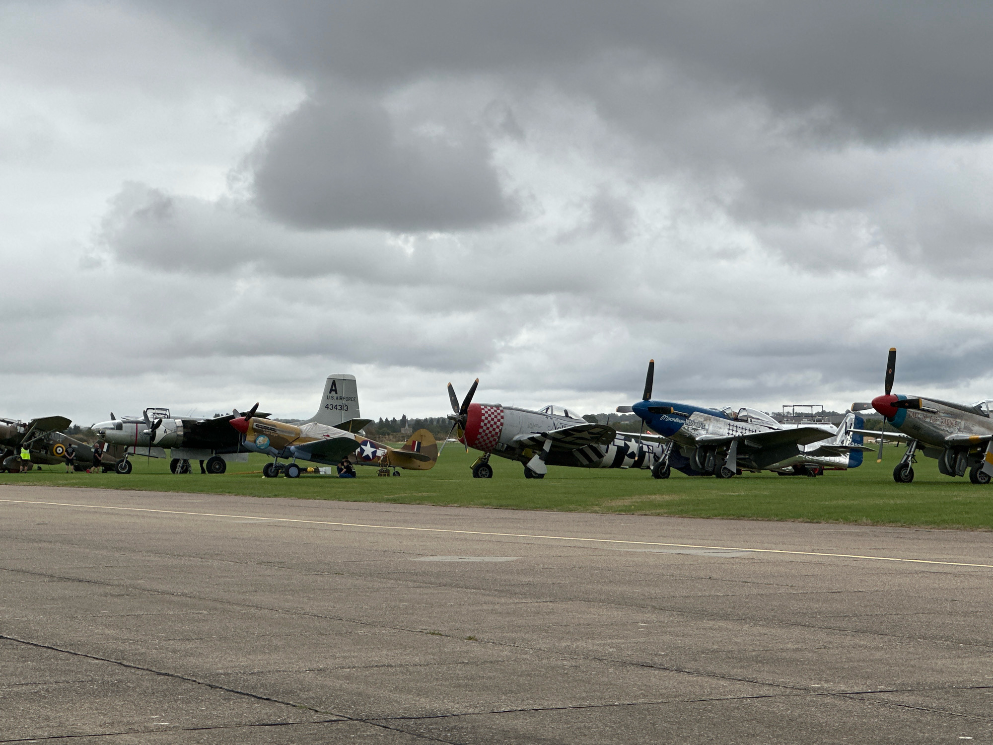The image depicts an outdoor scene at an airfield, likely a static display of vintage aircraft under a cloudy sky. The foreground shows a paved runway or tarmac, with faint white lines marking its surface. In the middle ground, a row of several historical airplanes are parked on a grassy area.
From left to right, the first visible aircraft appears to be a dark-colored propeller plane with circular insignia on its wings. Next to it is a lighter-colored twin-engine bomber with a distinctive nose, identified by the marking "A U.S. AIR FORCE 434313" on its tail. To the right of the bomber is a yellow and brown propeller plane with a star insignia, possibly a Curtiss P-40 Warhawk, followed by a red and white checkered-nosed aircraft, likely a Republic P-47 Thunderbolt. Further to the right are two blue and silver fighter planes, one with the name "Moonbeam" visible, and another with "In The Saude" written on its side. These appear to be North American P-51 Mustangs.
Several individuals are present, mostly seated or standing near the aircraft, suggesting they are either ground crew, enthusiasts, or visitors to an airshow. The overall environment is that of an airfield with a display of vintage military aircraft. There is no direct indication of the city of Cambridge, United Kingdom, within the image itself, but the presence of military aircraft and an airfield could be consistent with locations in the wider Cambridge area known for aviation heritage or events.