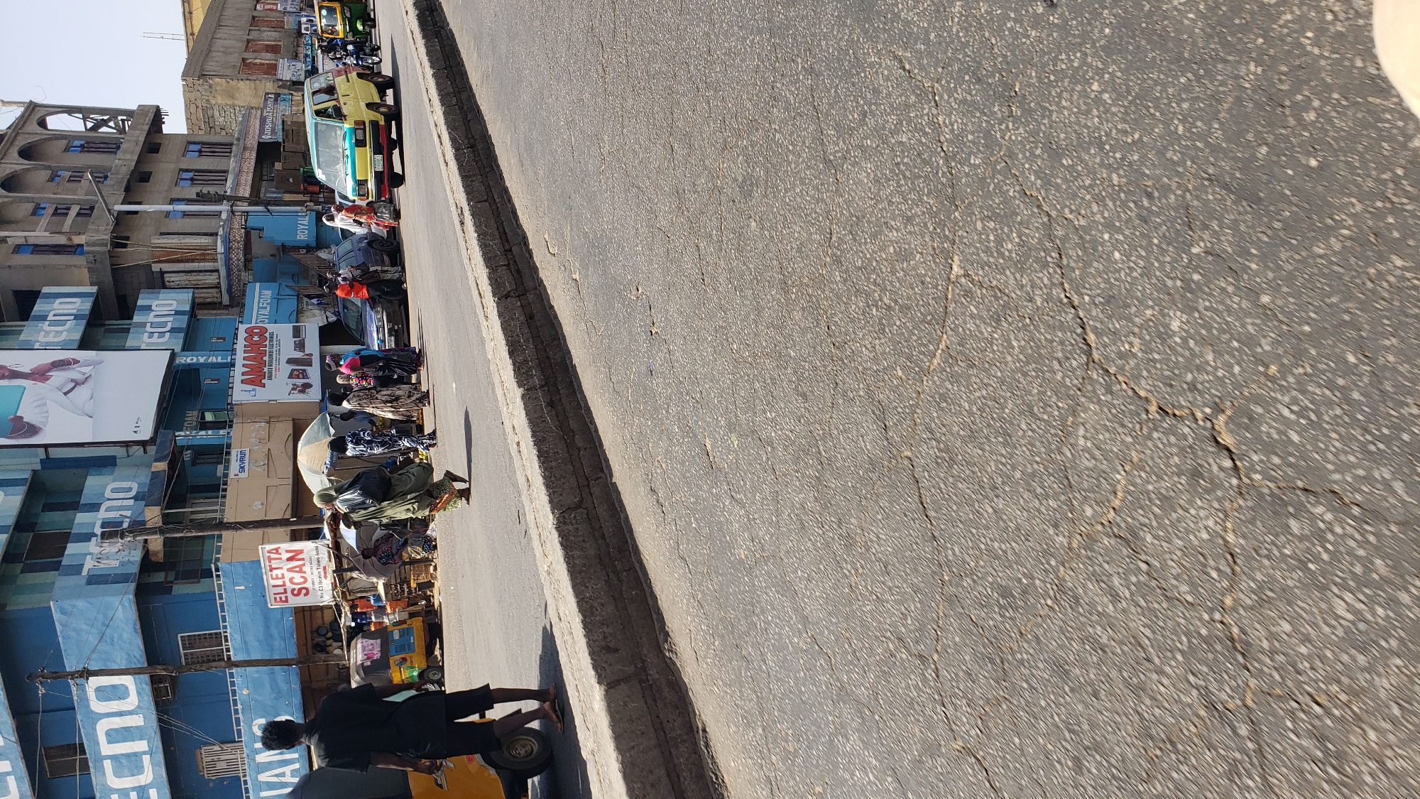The image depicts a street scene in Maiburiji, Nigeria. A cracked asphalt road dominates the foreground. Along the left side of the frame, buildings are visible, adorned with large advertisements for brands like TECNO, AMAHCO SYALFOAM, SKYRUN, and ELLETTA SCAN. People, including a man on a yellow motor tricycle (auto rickshaw), are observed near the buildings. Other vehicles, including a car and van, are parked nearby. The buildings are multiple stories high and of varying architectural styles, with some showing signs of age and wear.