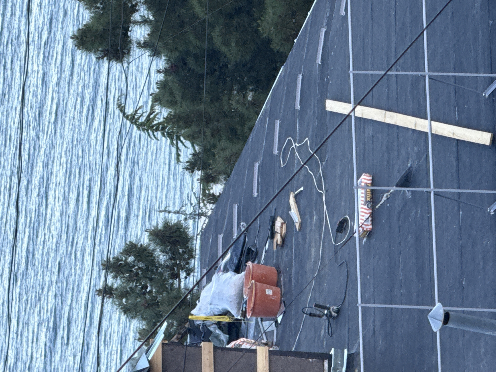 The image depicts a construction site adjacent to a body of water, possibly a lake or ocean. The site shows a roof under construction, evident by the black roofing material laid down, along with wooden beams, metal frames for solar panels, and various tools and equipment. There are orange buckets, power tools, and a white cable strewn across the roofing surface. In the background, there's a view of a water body, with trees visible near the construction site. The city and state remain unknown.