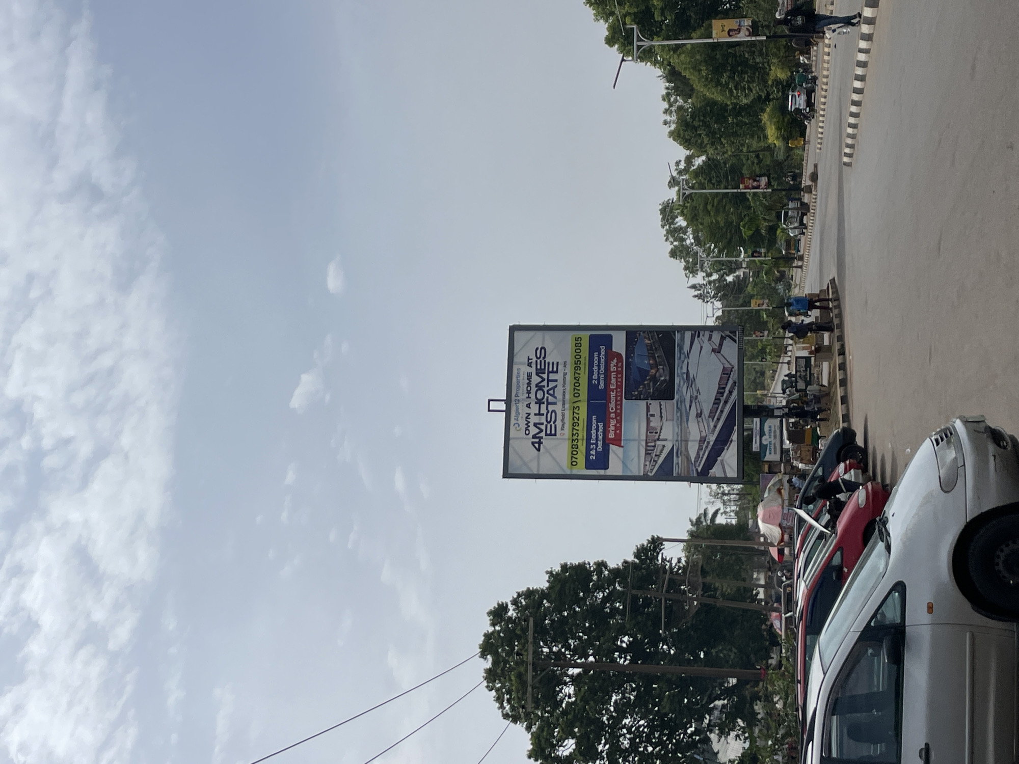 The image depicts a street scene in Jos, Nigeria. A large billboard advertising "4M Homes Estate" in Rayfield Extension is prominent. The billboard lists contact numbers and features information about 2 & 3-bedroom detached and semi-detached homes. Parked cars line the roadside. Trees are visible, and there are utility poles with power lines. People can be seen in the distance on the sidewalk. The sky is overcast.