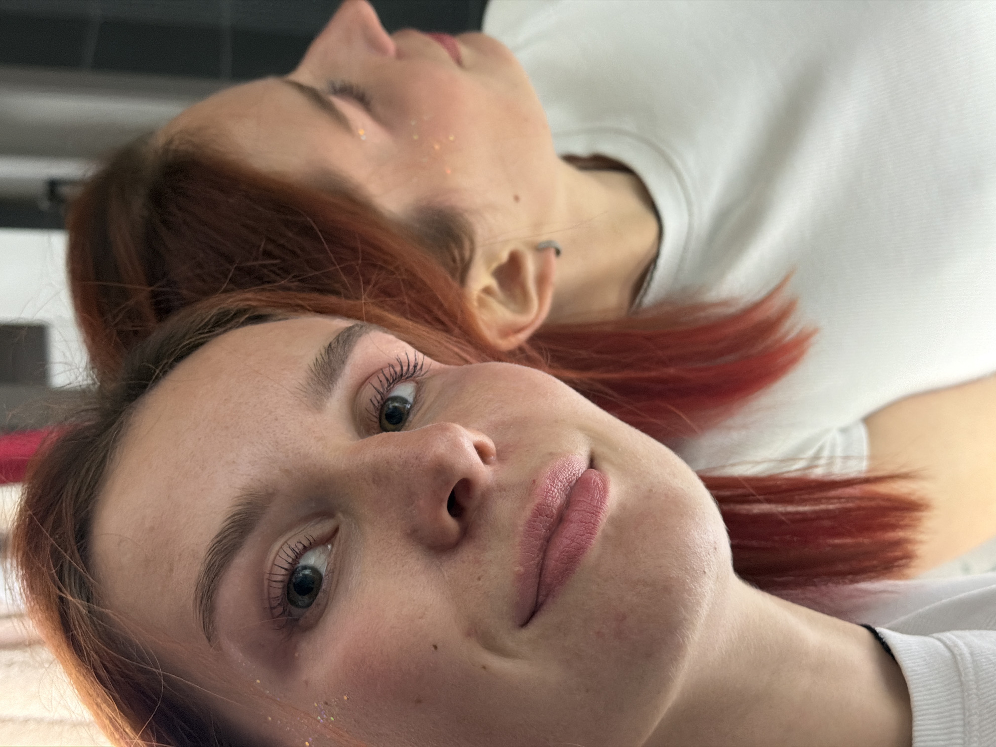 Two women with reddish-brown hair are lying down, possibly on a bed or flat surface, as suggested by the lighting and close proximity. The woman in the foreground is looking directly at the camera. There are sparkly spots on her cheek. Her hair is reddish-brown. The second woman is lying behind her, with her head tilted slightly upwards and her eyes closed. She also has sparkly spots on her cheek. Both women are wearing white t-shirts. The background is dark and indeterminate. There is no evidence within the image to suggest that this image was taken in the city of Chaikovsky, Russia.