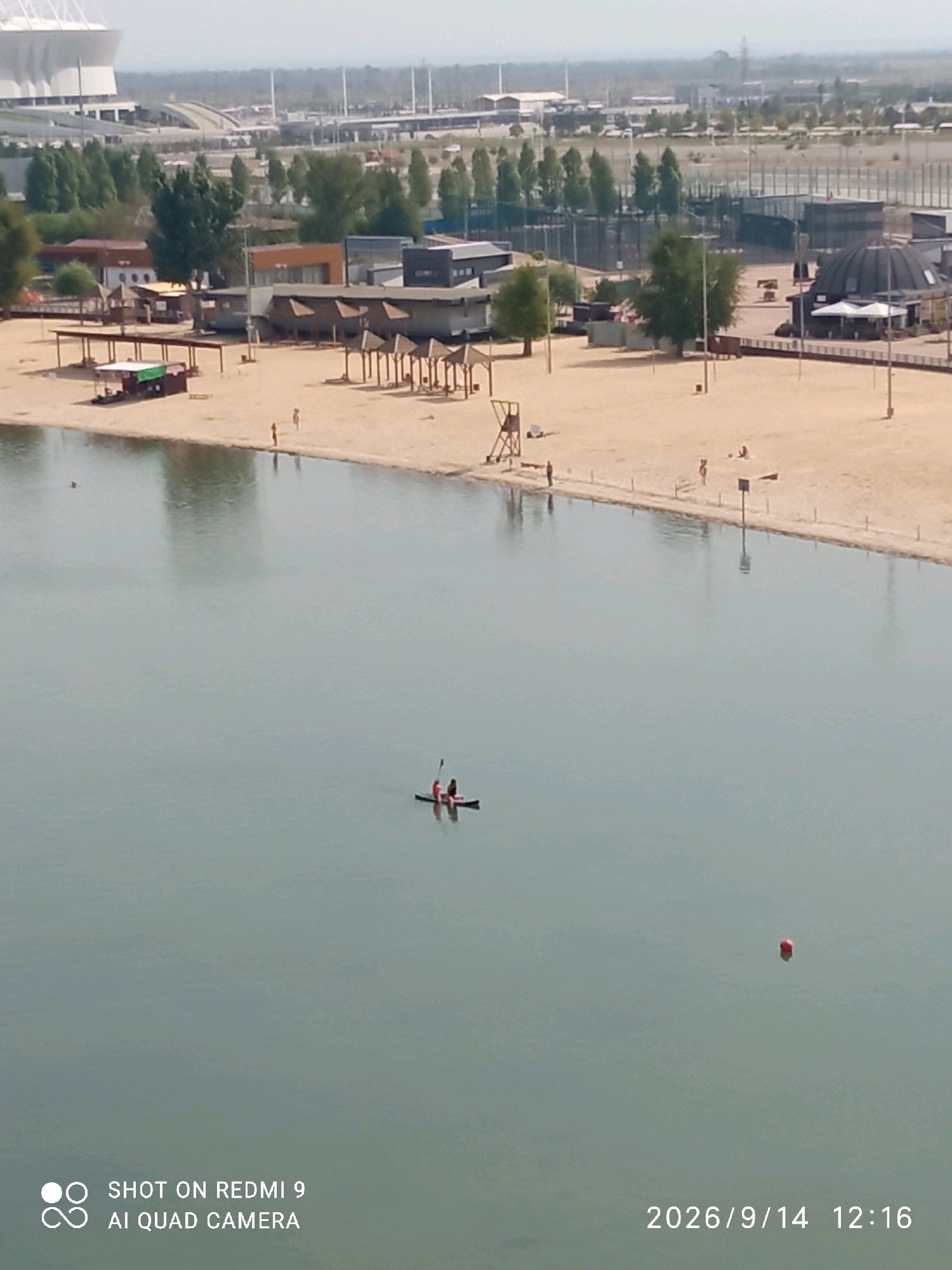 The image captures a serene scene at the Rostov Arena beach on a clear day. In the foreground, a tranquil body of water reflects the overcast sky. Two individuals are seen paddling a kayak on the water. The background reveals a sandy beach dotted with sunbathers and beach umbrellas. Further in the distance, the impressive architecture of the Rostov Arena stadium stands as a prominent landmark, with its distinctive white roof visible against the horizon. The overall atmosphere is one of leisure and relaxation, with the beach serving as a popular recreational spot within the city.