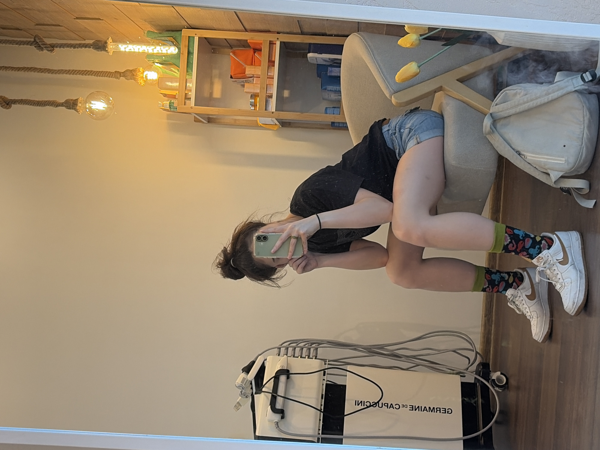 A person is taking a selfie in a mirror. They are wearing a black t-shirt, denim shorts, colorful socks, and white Nike Air Force 1 sneakers. The person is sitting on a chair with a gray seat and wooden legs. Next to the chair is a light gray backpack. In the background, there is a wooden shelf with various items on it, including orange boxes and blue containers. There are also several light bulbs hanging from ropes, which appear to be decorative lighting. In the foreground, a white machine with multiple cords is visible. The machine has text on it that reads "GERMAINE DE CAPUCCINI". The scene appears to be indoors, possibly in a room with light-colored walls and a wooden floor. The lighting suggests it could be daytime, or artificial lighting is being used. The person's posture and the selfie suggest a casual and relaxed moment.