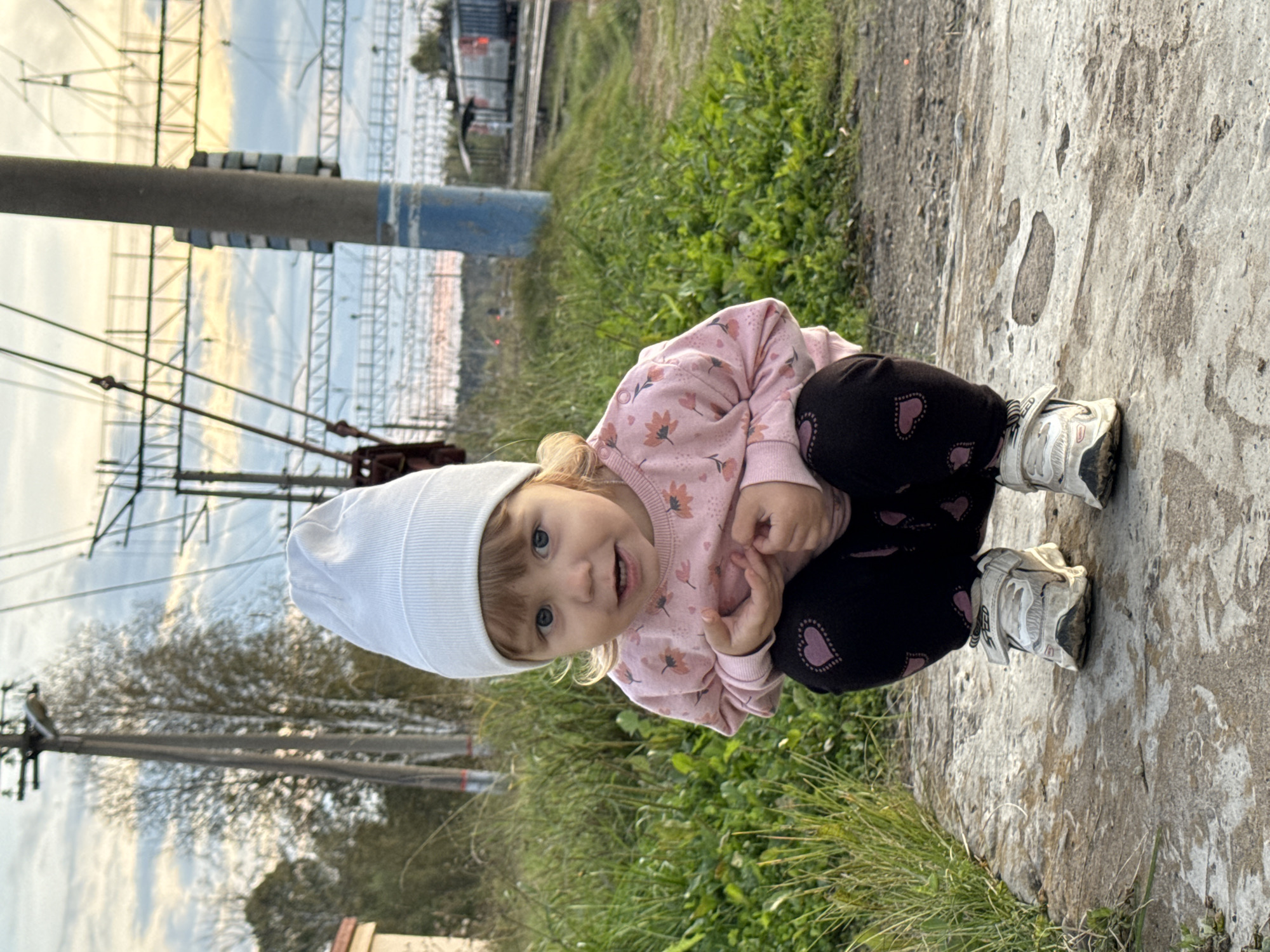 A young child with blonde hair and blue eyes is crouching on a concrete surface outdoors. The child is wearing a white knit hat, a pink long-sleeved shirt with floral patterns, and black leggings with pink heart appliques. The child's hands are clasped in front of their chest. The background shows green grass, trees, power lines, and a railway track with a train. The scene appears to be in or near Комарихинский, Russia. The lighting suggests it is late afternoon or early evening.