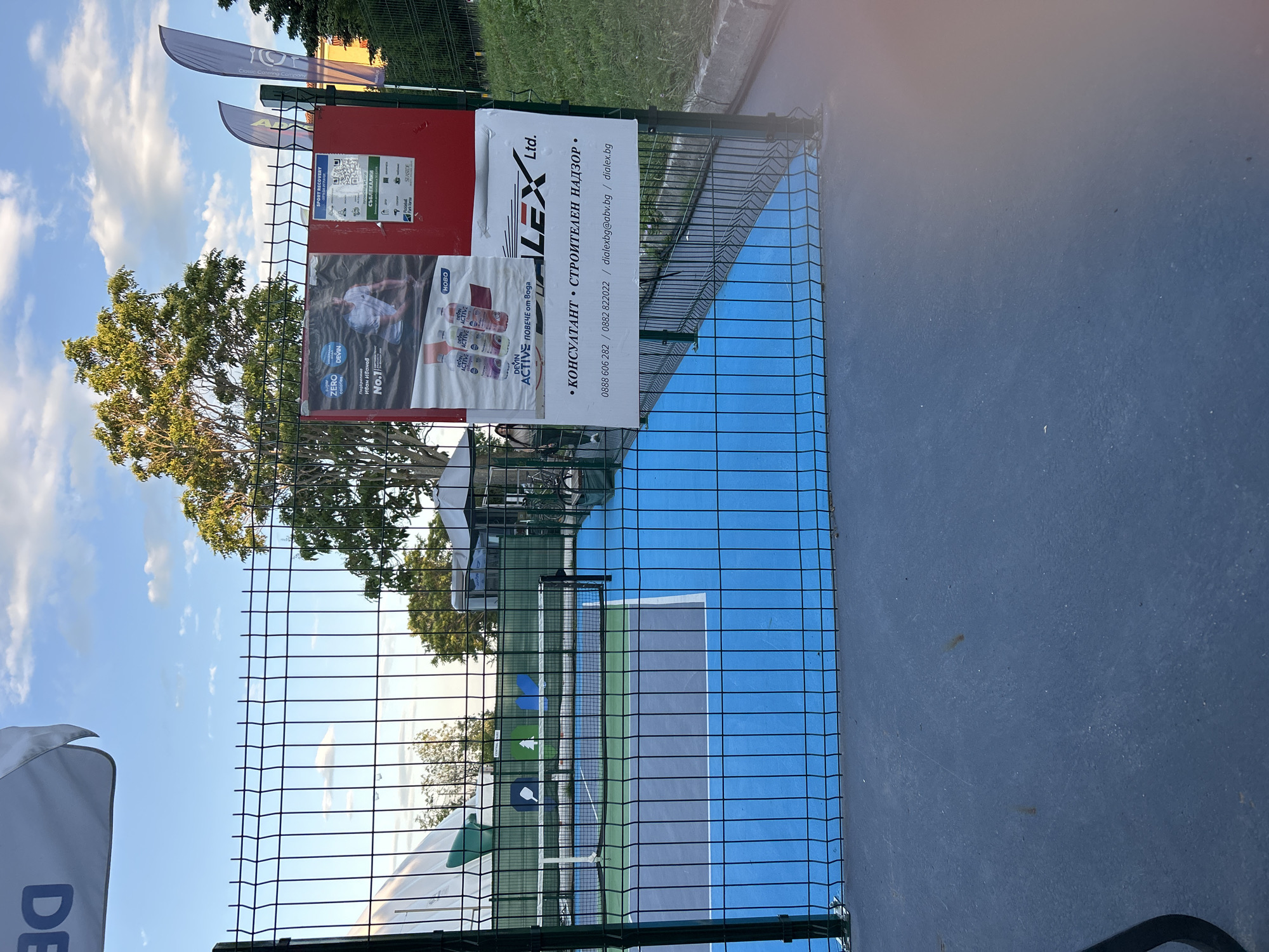 The image depicts an outdoor pickleball court environment located in Varna, Bulgaria, viewed through a dark metal mesh fence. The scene is illuminated by natural daylight under a blue sky with scattered white cumulus clouds.
In the immediate foreground, a dark grey paved surface, possibly asphalt or concrete, is visible. The prominent feature in the mid-ground is a dark green or black welded wire mesh fence, which serves as a barrier separating the viewer from the sports court.
Through the fence, the pickleball court is visible, featuring a bright blue playing surface with a dark grey or green perimeter and white boundary lines. A white net stretches across the court. Behind the net, another dark green fence is present, adorned with three distinct stylized logos: a white pickleball paddle, a green tree, and a blue heart-shaped or leaf-like design, above text that appears to read "CENTRAL PARK".
To the right of the court, attached to the fence, is a large red rectangular sign panel. Affixed to this panel is a large paper poster advertising "DEVIN ACTIVE" water, showcasing bottles and featuring a male athlete identified as Ivan Ivanov, promoting "ZERO calories" and "повече от вода" (more than water). Adjacent to this poster on the red panel are additional details, including the text "SPORT RECOVERY," "СЪБЛЕКАЛНИ" (Changing Rooms), a QR code, and logos for "Pickleball Park Varna" and "SEASIDE." Below the red panel, a white banner is attached, displaying the name "ALEX Ltd." and services: "• КОНСУЛТАНТ • СТРОИТЕЛЕН НАДЗОР •" (Consultant • Construction Supervision), along with phone numbers (0888 606 282 / 0882 822022), an email (dialexbg@abv.bg), and a website (dialex.bg).
In the background, multiple large trees with green foliage are prominent, particularly one large tree to the left of the signage. White event tents or canopies are visible; one is partially seen on the far left, and another is to the right behind the fence. A partially visible female figure with dark hair stands near the right-hand tent. Bicycles are parked near this right-hand tent. Further in the background on the far right, a segment of a light-colored building, possibly yellow or orange, is discernable behind the trees. Two vertical feather-style flags, one purple-blue and one grey-white, are positioned to the right of the red sign.