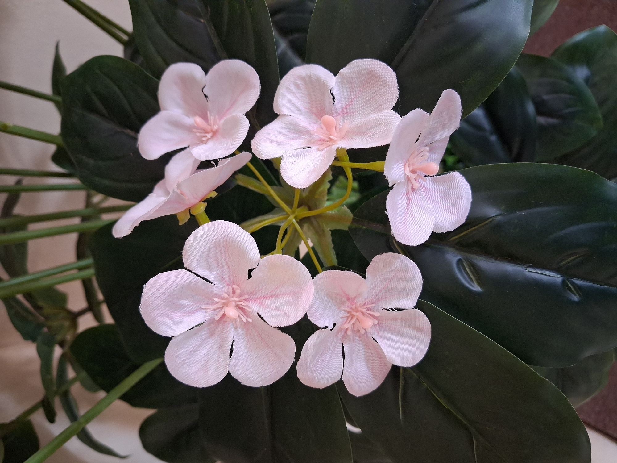 This image depicts a close-up view of artificial light pink flowers with visible stamens, set against a backdrop of dark green foliage. The flowers are arranged in a cluster, with five prominently displayed. The lighting suggests an indoor environment, possibly a home or a shop. The image's metadata does not contain information about its location in Bulungur, Uzbekistan, or any specific details about people or actions within the scene. The focus is solely on the artificial floral arrangement.