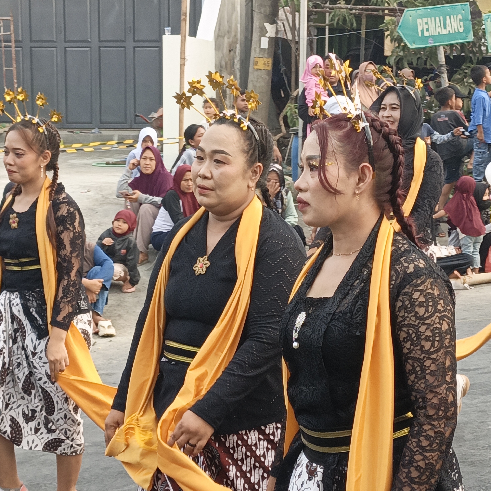 In Randudongkal, Indonesia, three women in traditional attire walk outdoors. The woman on the left wears a black lace top and a patterned skirt with an orange sash. Her hair is styled in braids and adorned with gold star-shaped ornaments. The central woman is dressed similarly, with a black lace top, patterned skirt, and orange sash, also wearing a floral brooch and gold star ornaments in her hair. The woman on the right, partially visible, wears a black lace top and an orange sash, with braids and gold star ornaments. A crowd of spectators, including children and adults wearing hijabs, watches them. The background features a gray building with dark doors and a green directional sign pointing to "PEMALANG." The ground is paved, and a yellow and black striped barrier is visible.