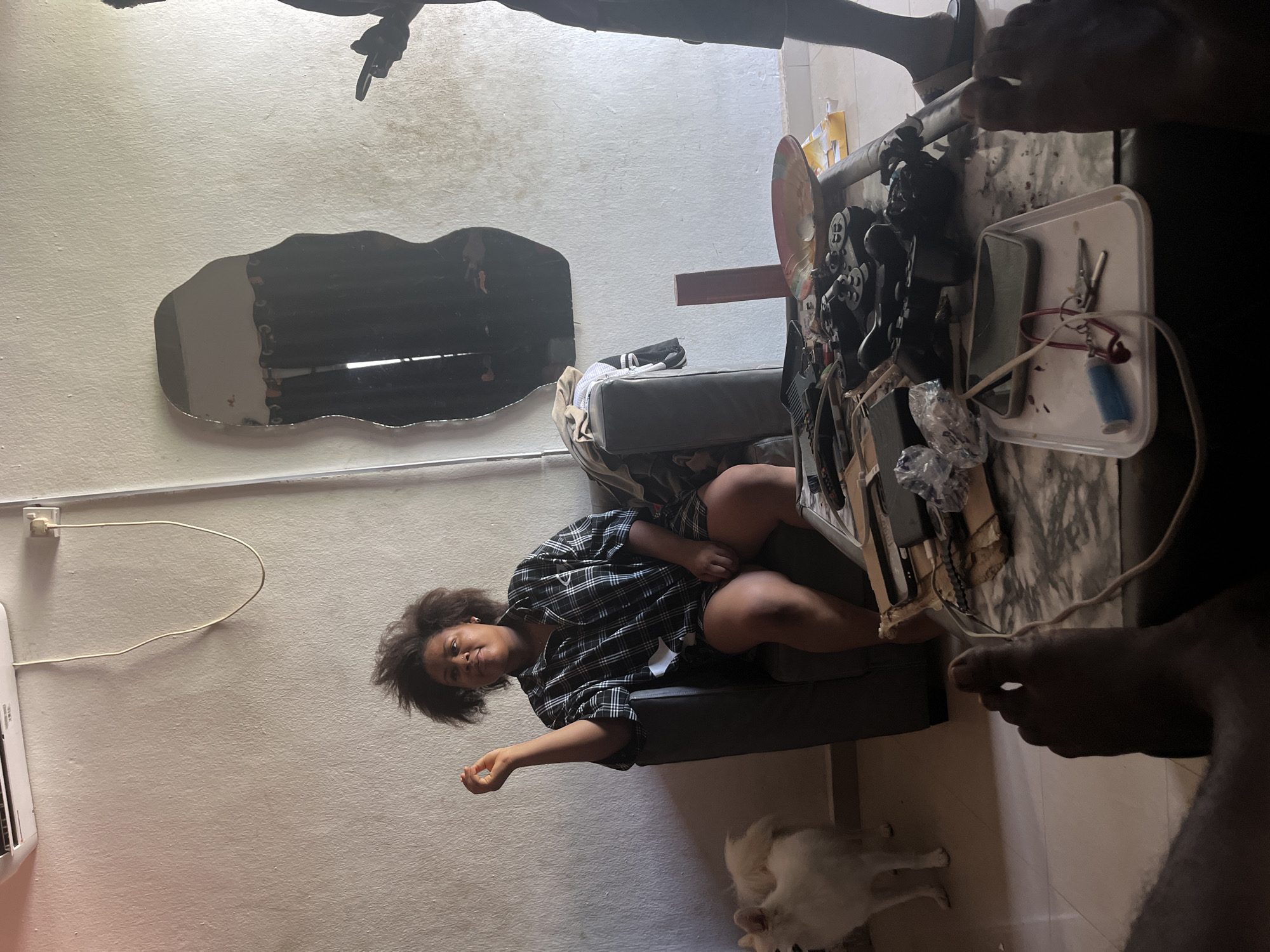 A room is seen in Ikot Ekpene, Nigeria. A woman sits on a grey chair wearing a plaid shirt. Next to her is a white dog. On a table are game controllers and a white tray with tools. A mirror hangs on the wall behind the chair. Cables run along the wall. Several people are partially visible near the table.