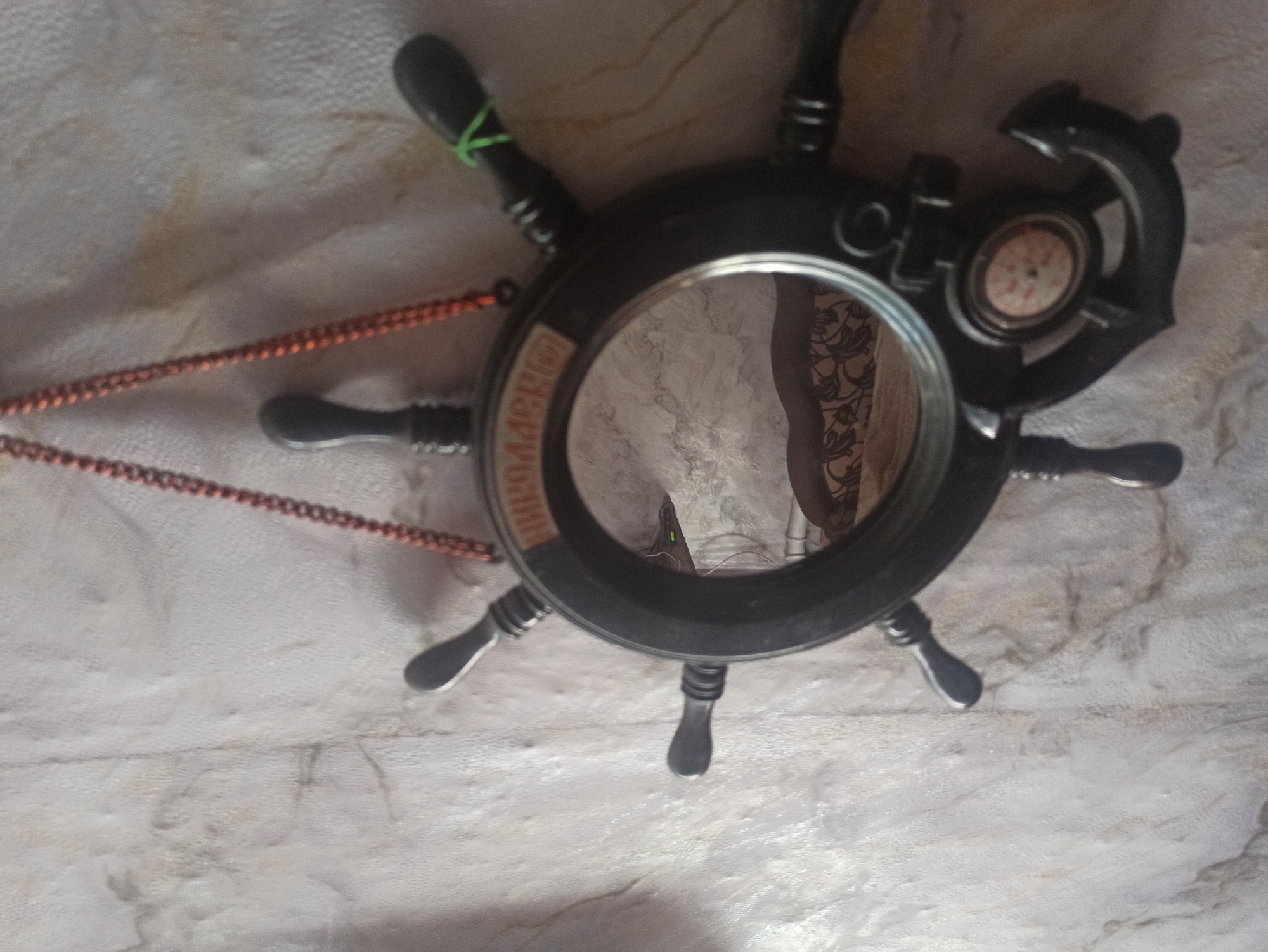 The image shows a decorative nautical-themed mirror, shaped like a ship's steering wheel with an anchor at the bottom. The mirror is hanging on a textured, light-colored wall with a marble-like pattern. The steering wheel is black with several spokes that have small handles. A thin copper chain is visible at the top, suspending the decoration. Inside the steering wheel, a mirror reflects a dimly lit room. The reflection shows a pattern on what appears to be upholstery or fabric, possibly a sofa or bed, and a digital clock displaying "11:00". There is no visible connection to the city of Kryvyi Rih, Ukraine. The object appears to be indoors.