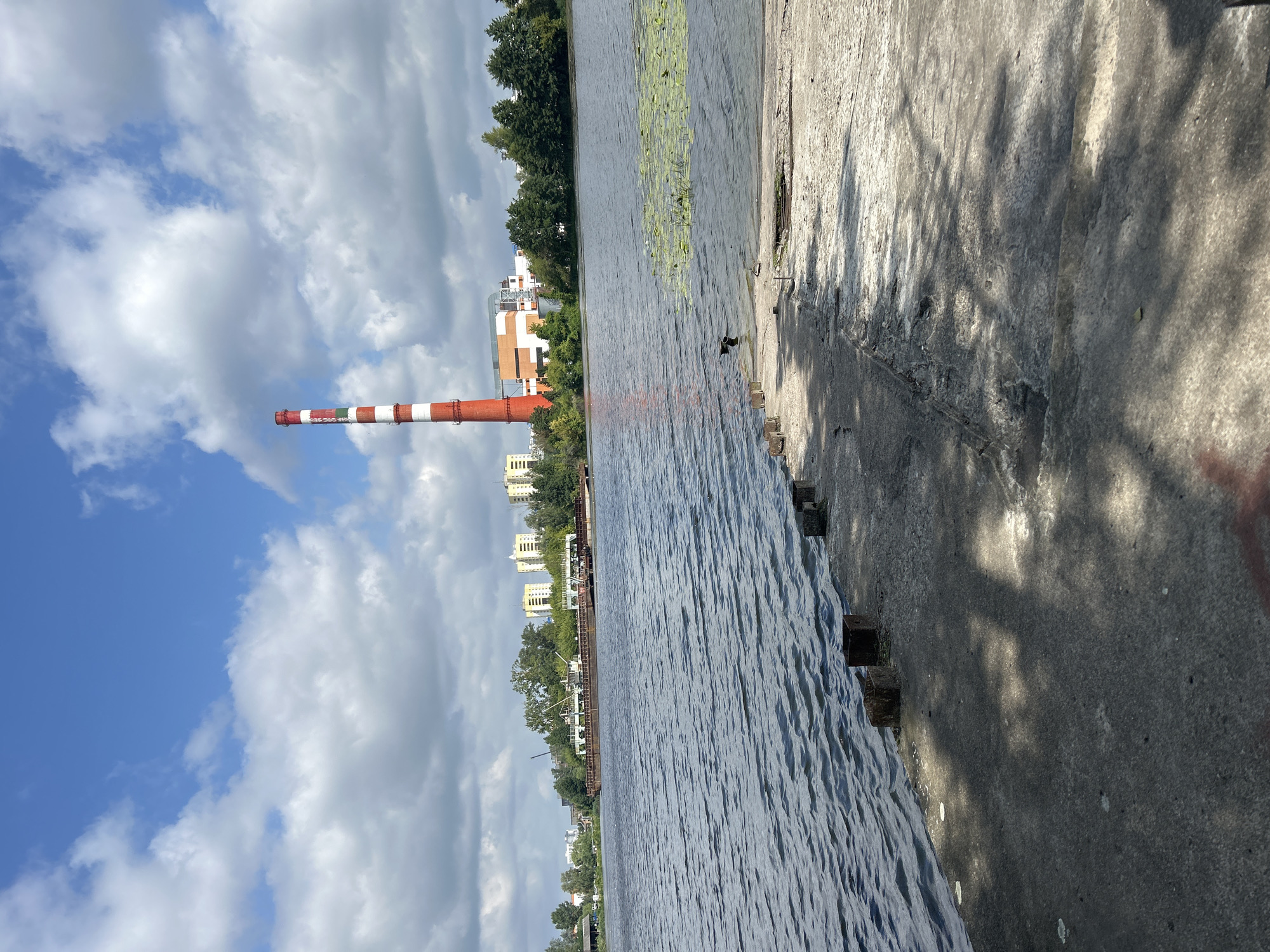 An outdoor, full shot captures a wide, rippling body of water, likely a river or canal, under a partly cloudy sky. In the foreground, a weathered, gray concrete pier juts into the water, with several rusty, square metal posts protruding from its surface. The water reflects the sky, creating a textured, shimmering effect.
Across the water, the opposite bank is lined with dense green trees and several multi-story buildings. Prominently featured on the right side of the bank is a tall, red and white striped industrial chimney, a characteristic landmark of Brest, Belarus. To the left of the chimney, an industrial building complex is visible, with orange and white structures. A long, flat-bottomed barge with cranes is moored near the shore, with more industrial buildings and trees behind it. Two black birds are visible in the water, near a patch of lily pads. The overall atmosphere is one of industrial activity and a partly overcast summer day.