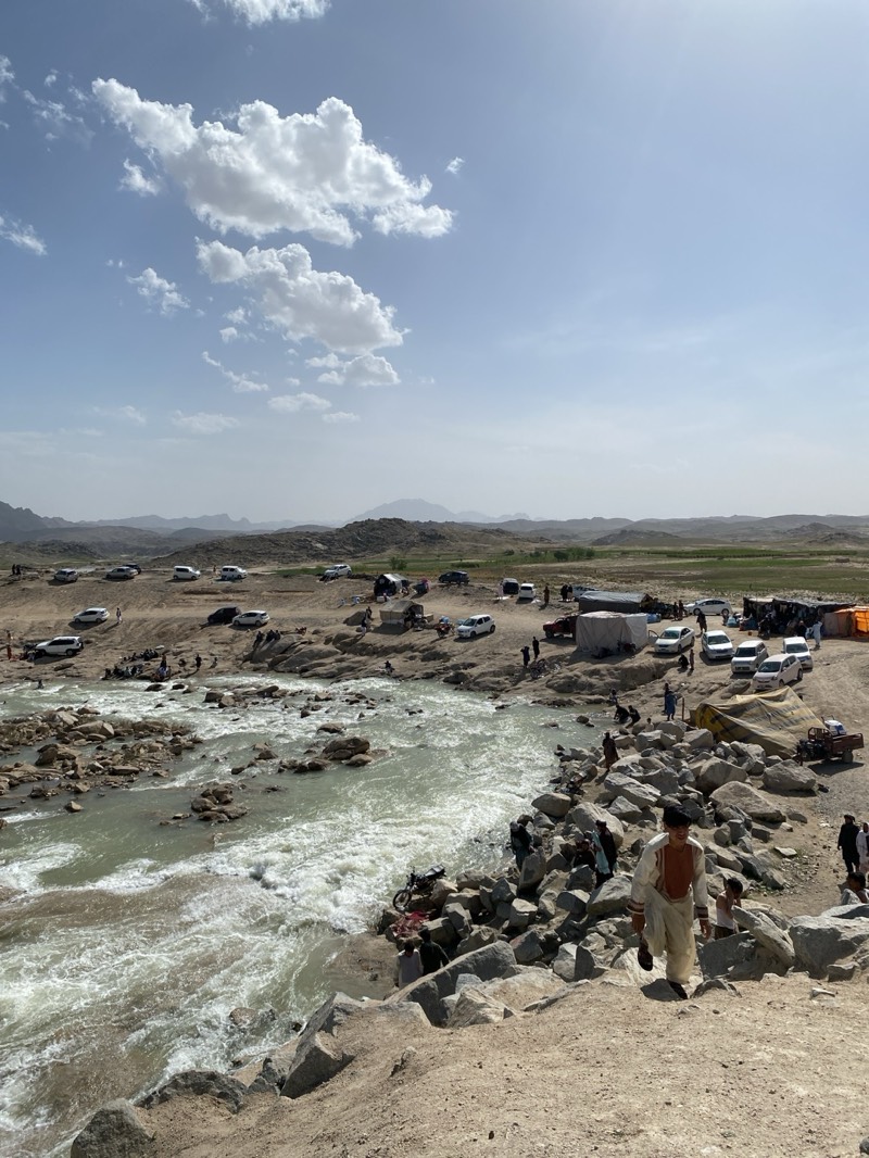 The image depicts a vibrant scene in Kim, Afghanistan, showcasing a river flowing through a rocky landscape. The river's waters are turbid, suggesting a strong current. On both banks of the river, people are gathered, some in the water, others on the rocks or the shore. Several vehicles are parked nearby, indicating a popular gathering spot. Tents and shelters are erected on the land, possibly for picnics or gatherings. One individual is seen walking down a rocky embankment towards the river, wearing traditional clothing. The sky is partly cloudy, with visible mountain ranges in the distance. The overall environment suggests a natural outdoor recreational area.