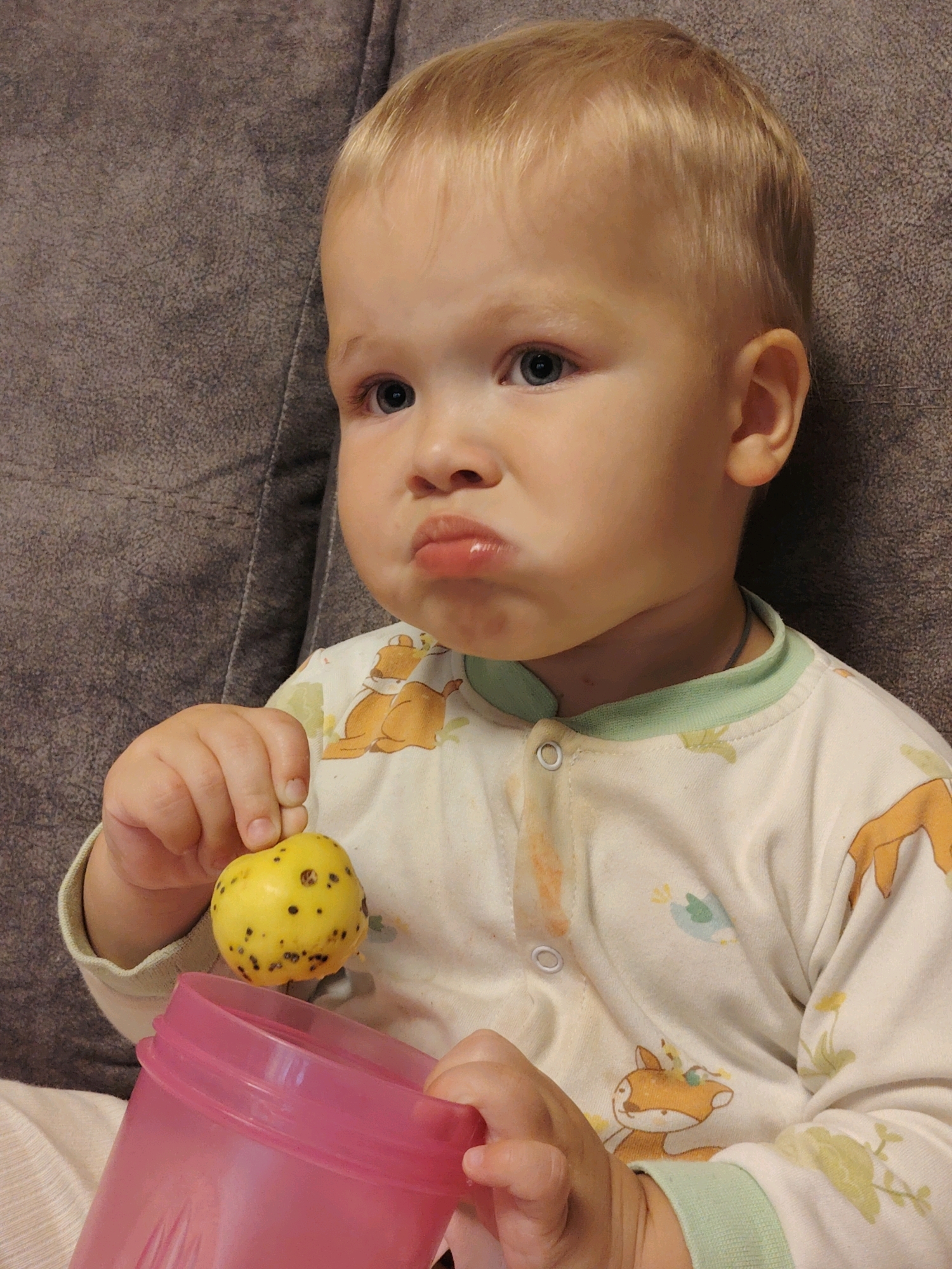 A close-up, eye-level shot shows a young child with blond hair and blue eyes sitting on a gray textured couch. The child is wearing a light-colored pajama set with cartoon deer and bird prints, and the collar is a pale green. The child's lips are pursed. In the child's right hand, they are holding a small, yellow fruit with dark spots. In the lower part of the image, a pink plastic cup is visible. The background is the couch. The scene appears to be indoors, likely in a residential setting in Chaikovsky, Russia.