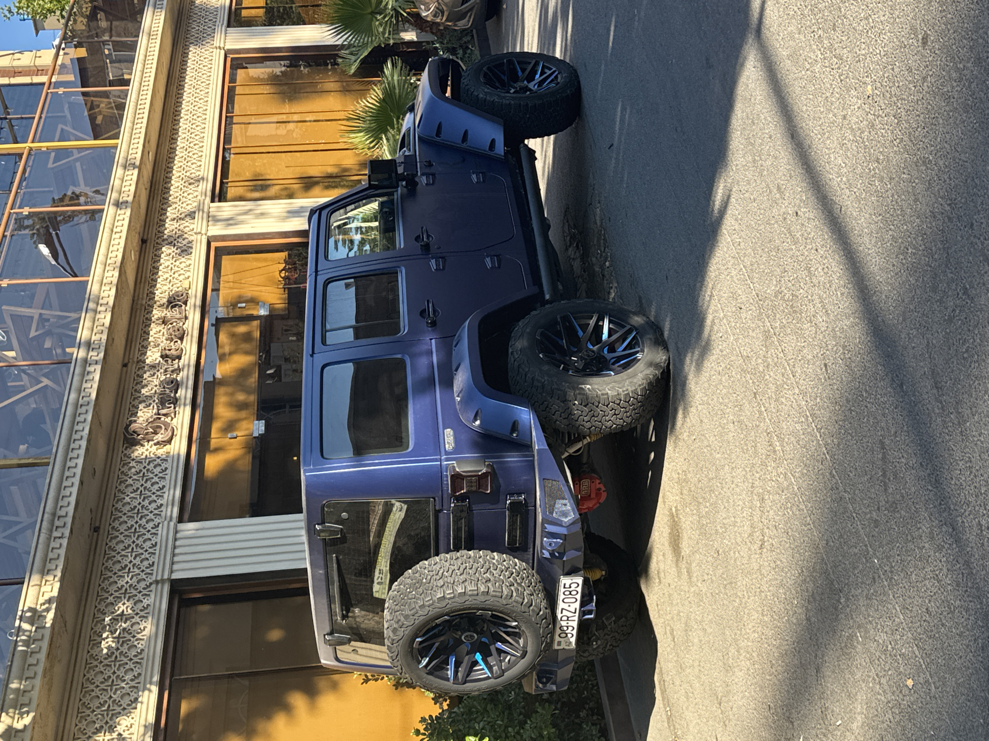 A navy blue Jeep Wrangler is parked on an asphalt street in front of a building with gold-tinted windows and ornate architectural details. The Jeep has a spare tire mounted on the back, custom wheels with blue accents, and a license plate reading "99-RZ-085". The building appears to be a commercial establishment, possibly a restaurant or cafe, with outdoor seating visible on an upper level. The scene is bathed in sunlight, casting shadows on the street and the facade of the building. Palm trees are visible on the right side of the image. The location is identified as Baku, Azerbaijan.