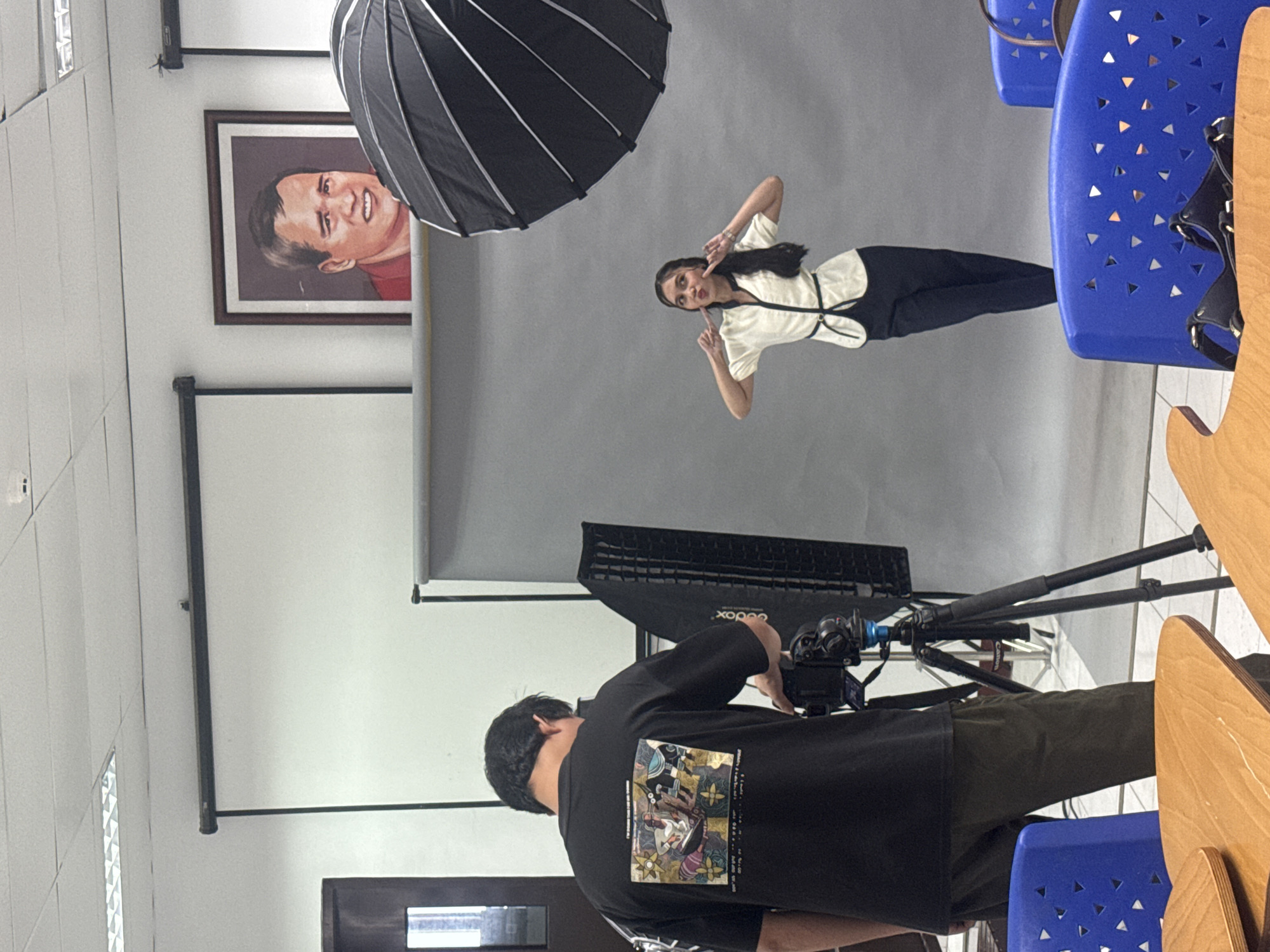 The image depicts a studio setup for a photo shoot. A person wearing a black t-shirt with a graphic print is operating a camera mounted on a tripod. To the right, a young woman in a white top and dark pants poses with her hands by her face, looking towards the camera. A large umbrella-style reflector is positioned above her. In the background, a grey backdrop is set up, and a framed portrait of a man is displayed on the wall. The environment appears to be indoors, possibly a classroom or a studio space, with fluorescent lighting visible on the ceiling. There are also blue chairs and wooden tables in the foreground, suggesting the setting could be an educational institution or a shared workspace. The location is identified as Davao City, Philippines.