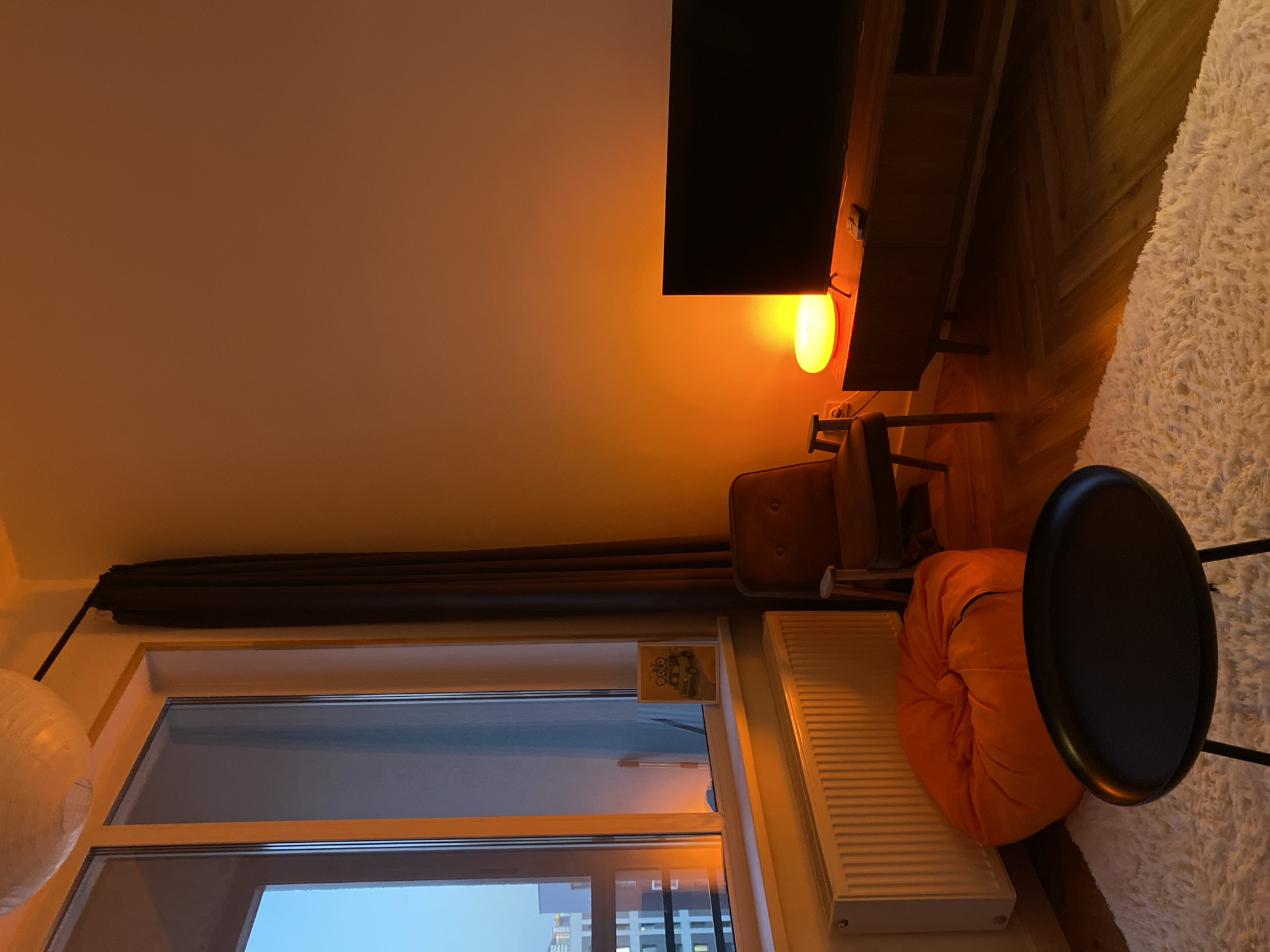 The image shows an interior scene featuring a window view of what appears to be Izhevsk, Russia. A curtain hangs to the left of the window, partially obscuring the view. A white spherical lamp is suspended near the curtain. The window frame is white. Outside, several buildings are visible beneath a blue sky.
Inside, a radiator is positioned near the window. To the right of the radiator is an orange beanbag chair. A black side table is placed next to the beanbag. An upholstered chair sits closer to a television stand, above which is a flat-screen television. The wall is painted in a light orange color. A lamp emits a warm orange glow. A fluffy rug is partially visible on the right side of the image. The floor is made of wooden planks.