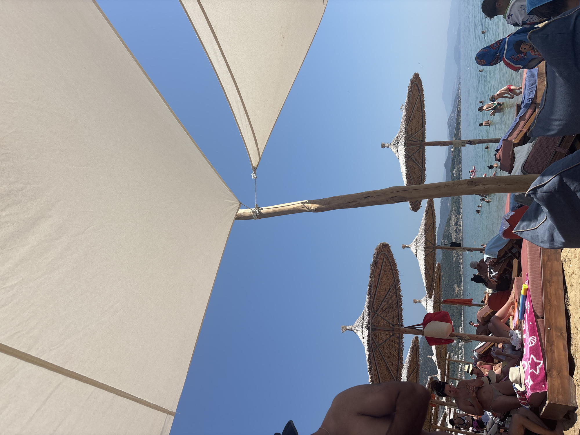The image depicts a crowded beach scene at Nea Iraklitsa, Greece, on a clear, sunny day. In the foreground, a large white canopy shades a portion of the scene, with a wooden support pole visible. Several straw-thatched umbrellas are set up in rows on the sandy beach, providing shade for lounge chairs occupied by beachgoers. In the middle ground, people are relaxing on the chairs, some reading, others on their phones, and a few are in the shallow water. The sea is calm and blue, with a distant coastline and hills visible under a clear sky. Various beach accessories, such as towels, bags, and a child's inflatable toy, are scattered around the lounge chairs. The overall impression is of a lively and popular beach destination.
