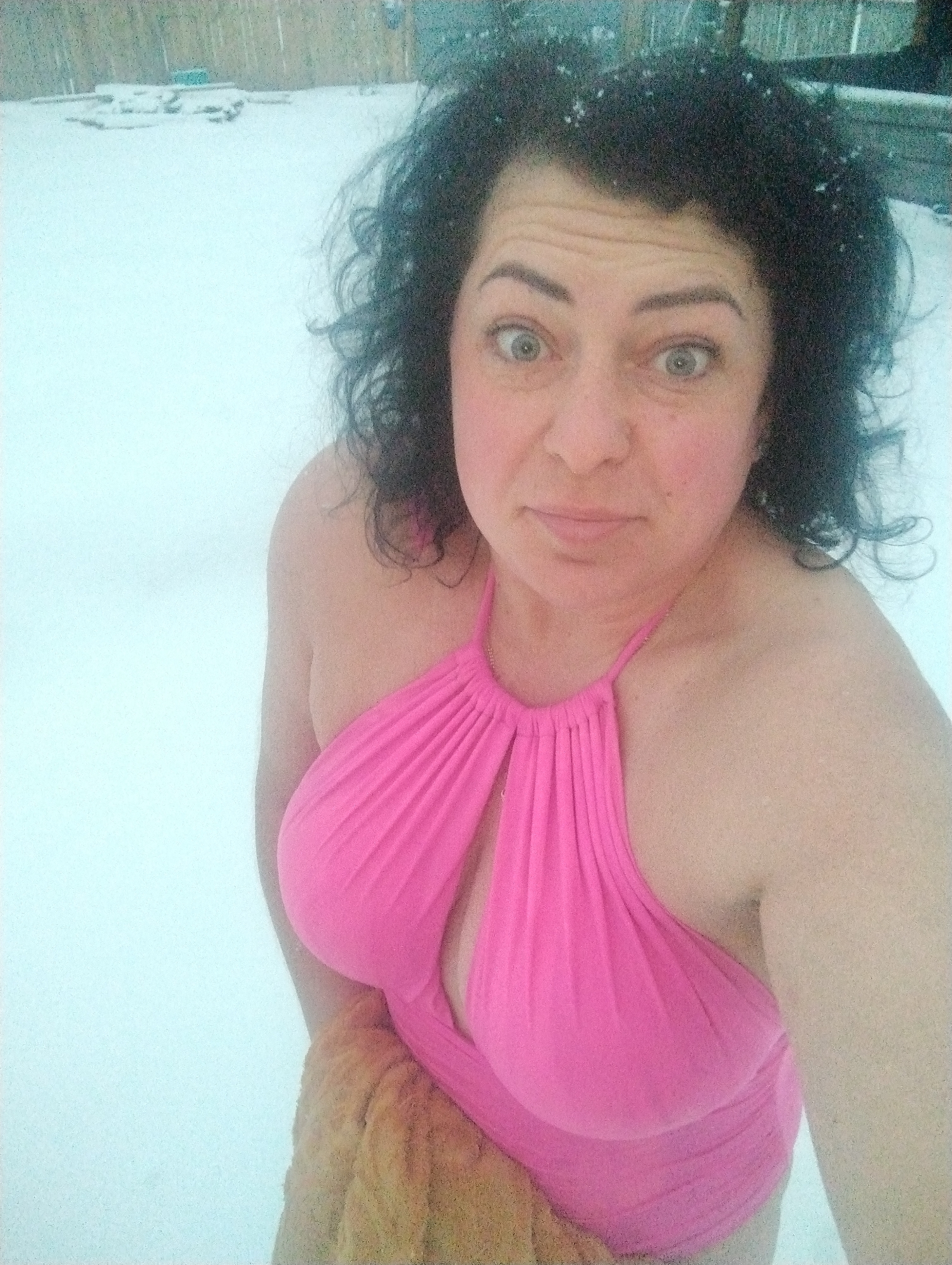 A woman with curly dark hair is standing in a snow-covered environment, possibly outdoors. Snowflakes are visible in her hair and on the ground. She is wearing a pink halter-style swimsuit and is partially covered by a tan-colored object. The woman's expression appears to be surprised, with wide eyes and a slightly pursed mouth. The location of Grishanki, Russia is not definitively evident in the image, but the snow and visible structures in the background could support this possibility.